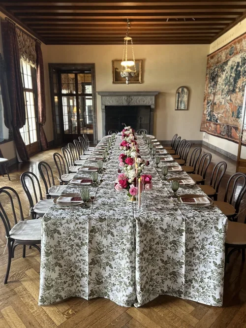 A long dining table set for a meal, with floral tablecloth, pink flowers centerpiece, and place settings with plates, glasses, and napkins. The room has wooden floors, large windows with curtains, a fireplace, and an ornate wall hanging.