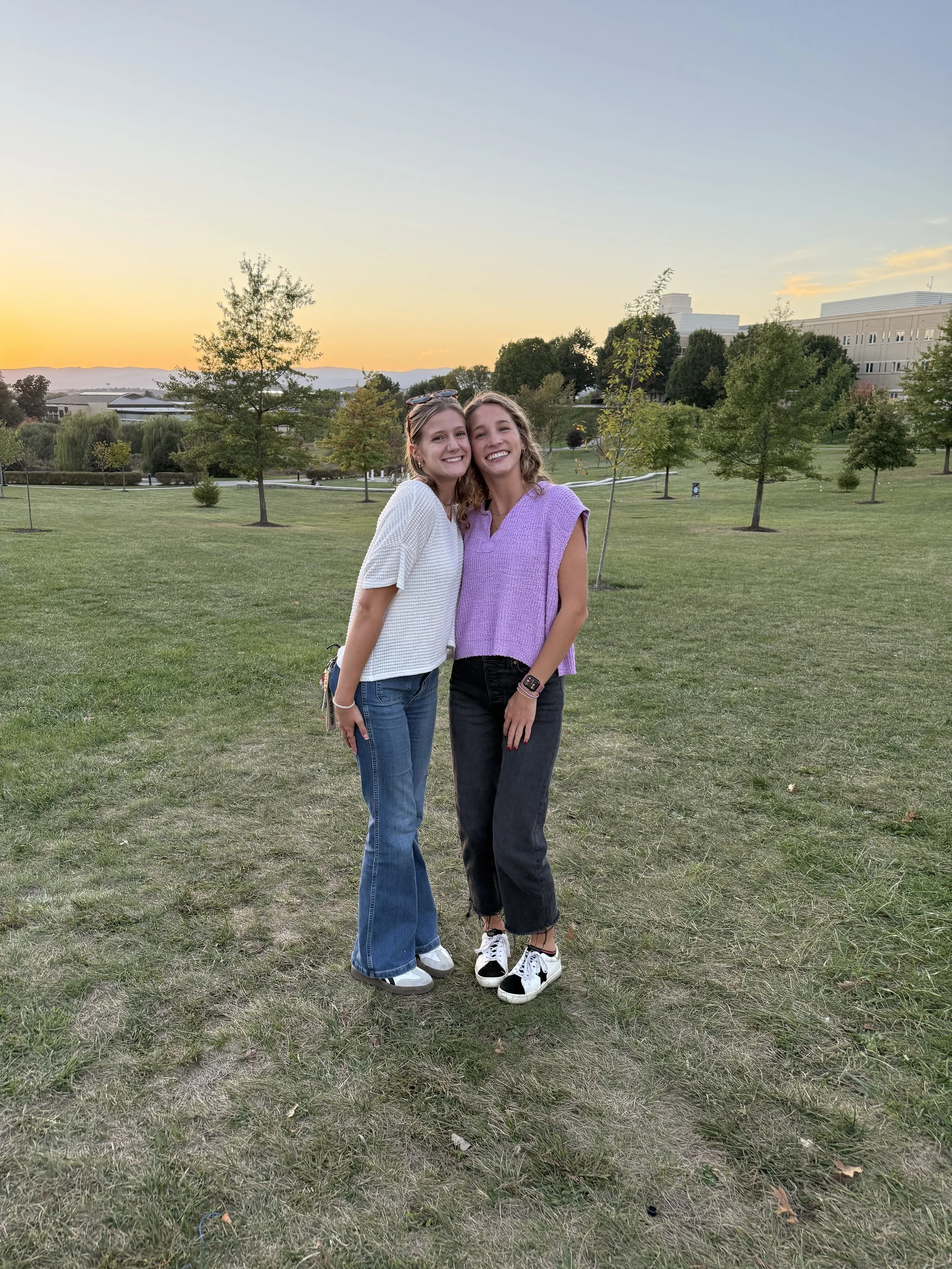 Two young women standing close together on a grassy park at sunset, smiling and looking friendly. One woman has blonde hair and wears a white top and jeans; the other has light brown hair, a purple sleeveless top, and black jeans, with sneakers. Trees and a large building are in the background.