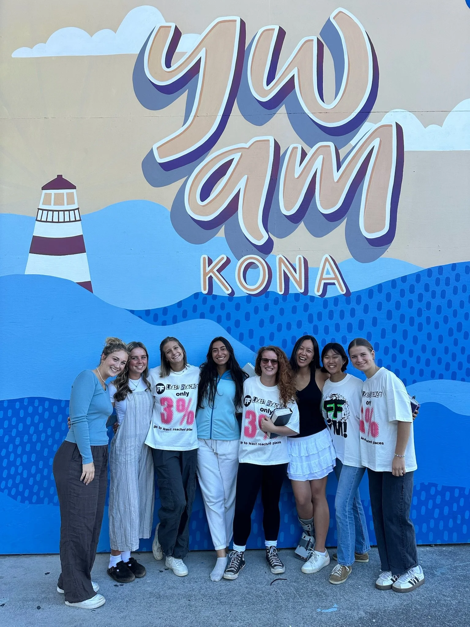 A group of nine young women standing in front of a blue mural with the words 'YJOY' and 'KONA' painted on it, smiling and posing for the camera.