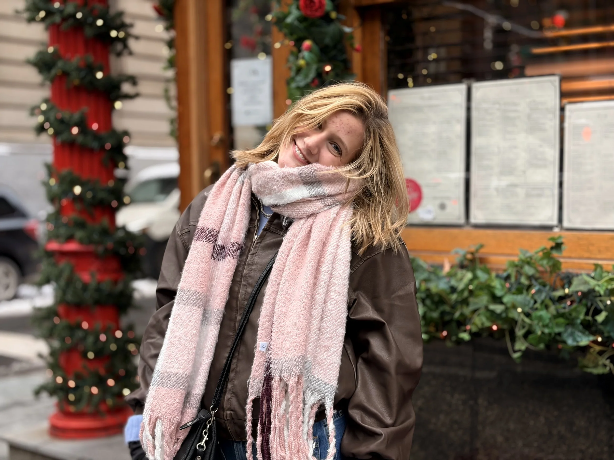A smiling young woman with long blonde hair, wearing a pink scarf and a brown jacket, standing outdoors during Christmas season with a decorated Christmas tree and holiday decorations in the background.