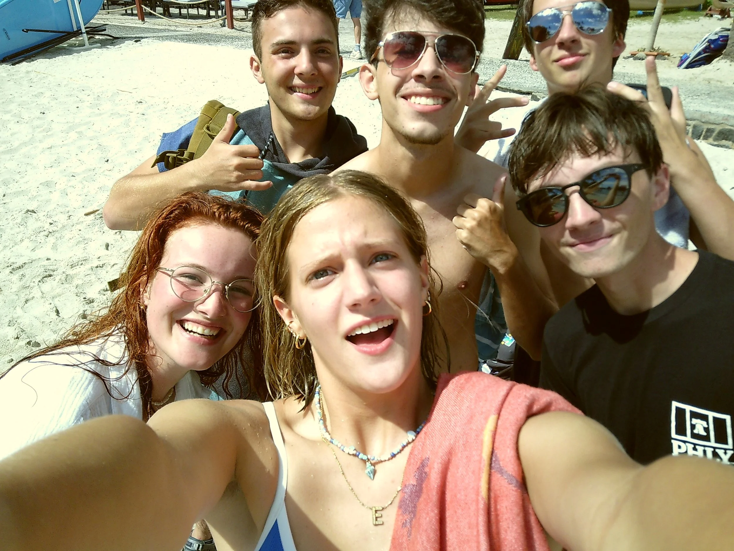 Group of five smiling young people taking a selfie on the beach, some wearing sunglasses, sand and beach equipment visible in the background.