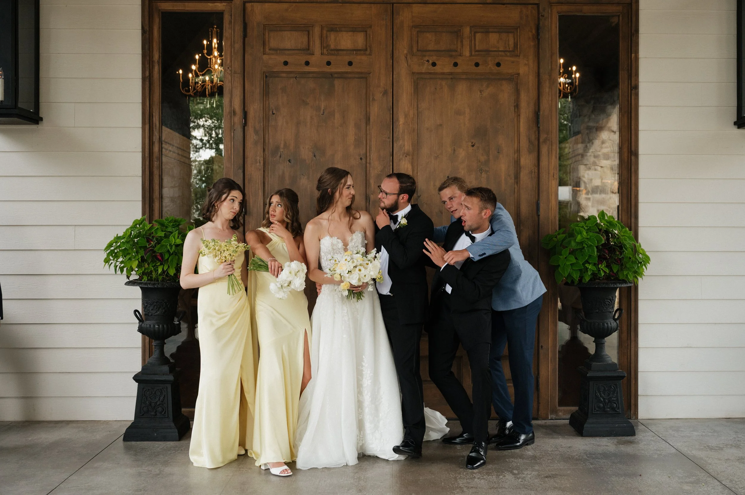 A group of six people at a wedding, including bride in a white gown, bridesmaids in yellow dresses, and groomsmen in tuxedos, standing in front of a large wooden door with two potted plants on either side.