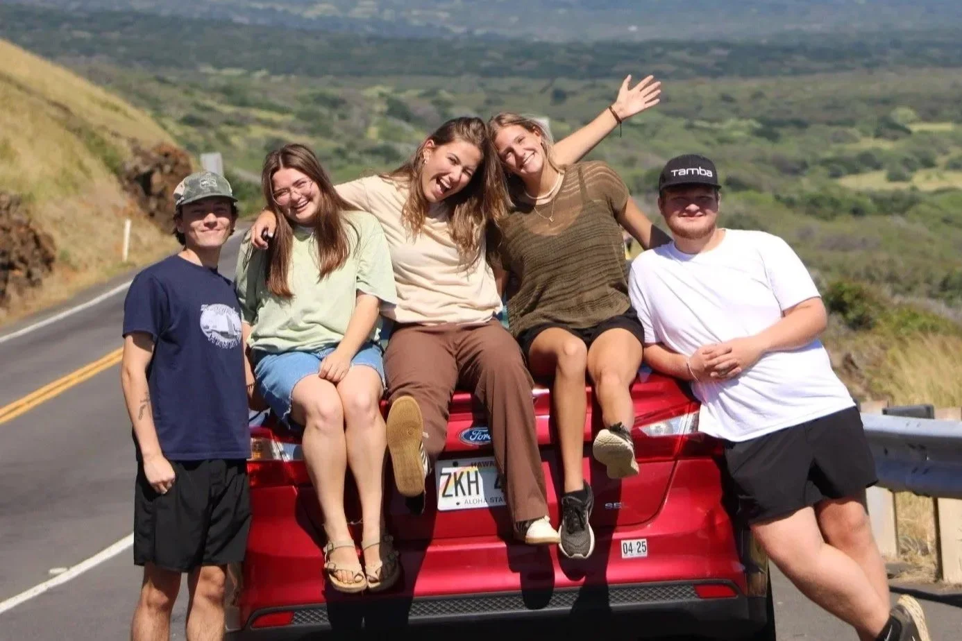 Five young people enjoying outdoors, sitting on the back of a red truck on a scenic highway with green hills in the background, smiling and waving.