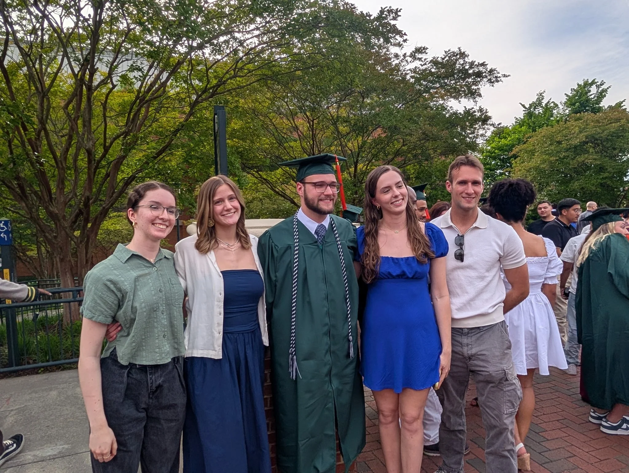 Group of five young adults, four women and one man, smiling and posing together outdoors during a graduation celebration. The man is wearing a cap and gown, while the women are casually dressed.