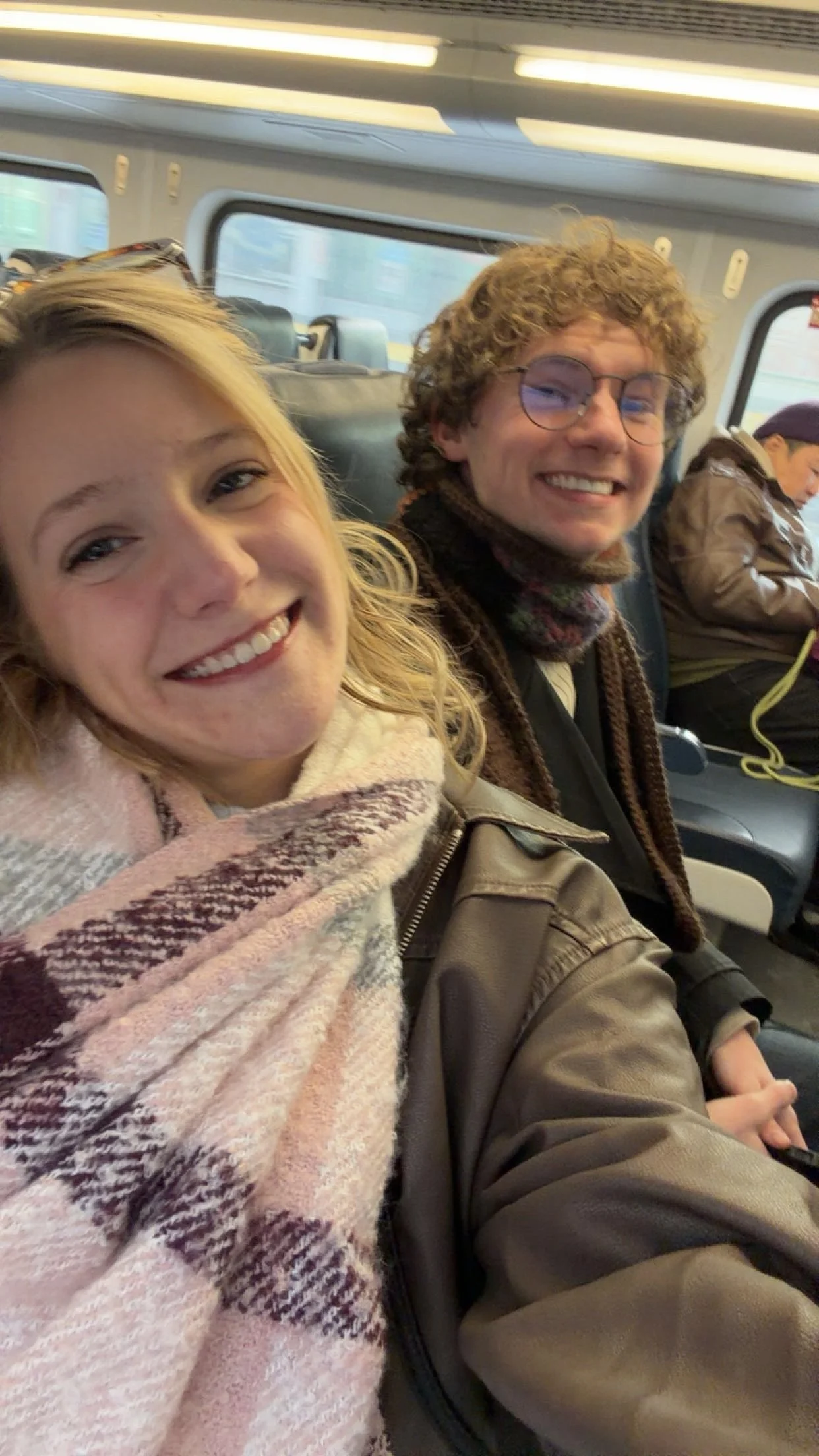 Two smiling people taking a selfie on a train, with a woman on the left and a man with curly hair and glasses on the right, and a sleeping person in the background.