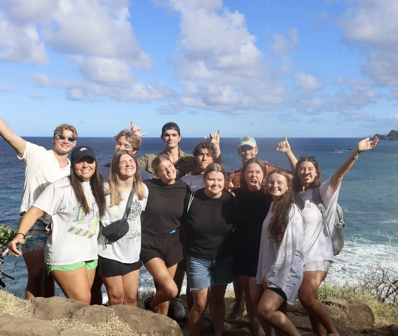 A group of twelve young people posing together on a rocky shoreline with the ocean and blue sky with scattered clouds in the background. They are smiling, wearing casual clothes, and some have hiking backpacks.
