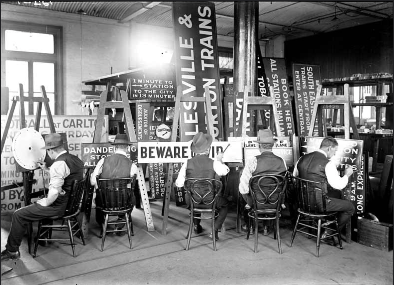 A vintage black and white photo of four men sitting on chairs at a table with signs and posters about transportation and warnings, inside a room filled with large signs and boards about train schedules and directions.