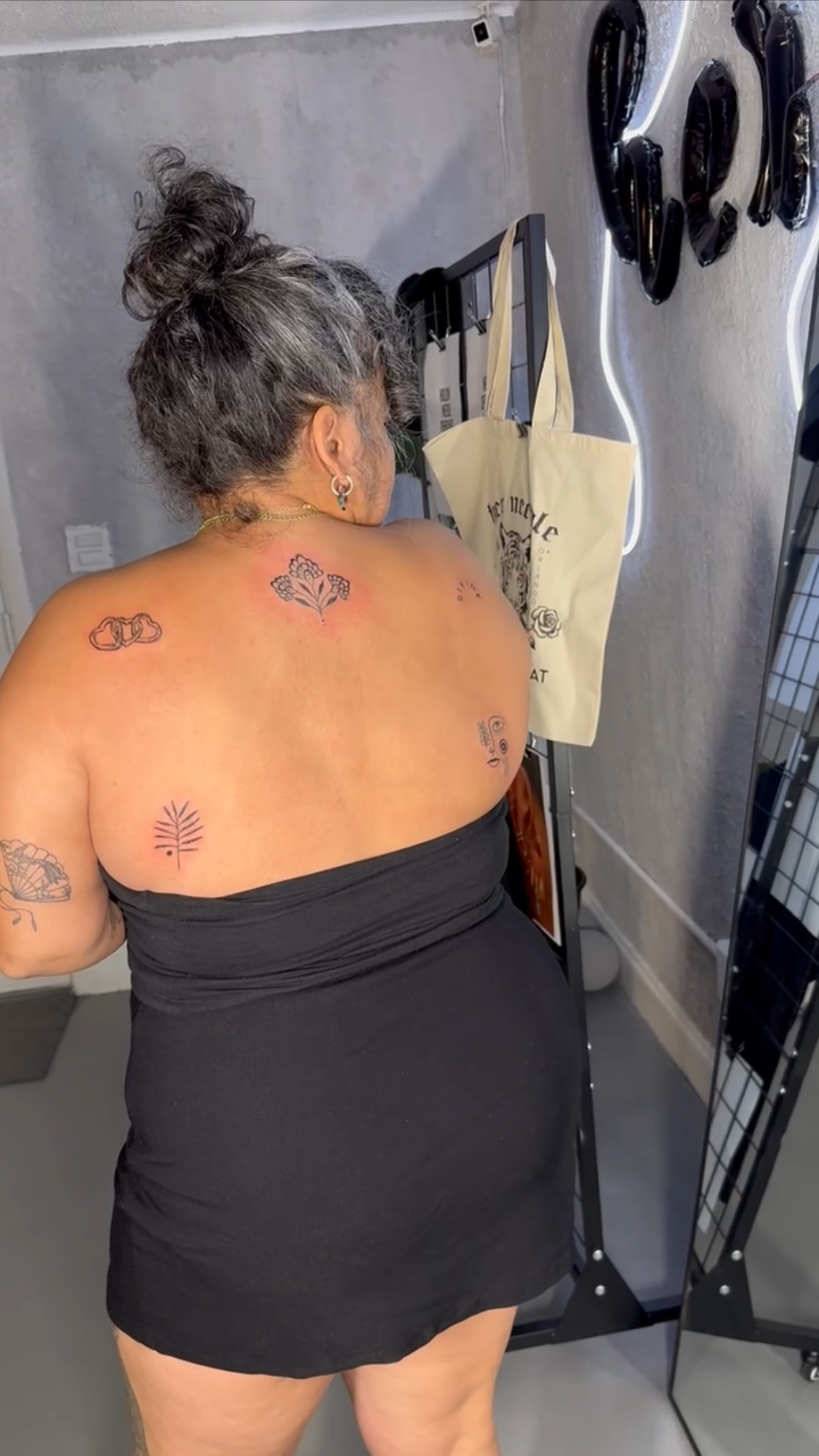 A woman with gray and black hair in a bun, wearing earrings and tattoos on her shoulder and back, is standing with her back to the camera in a room with gray walls and a gray floor. She is wearing a strapless black dress. There is a beige tote bag ha