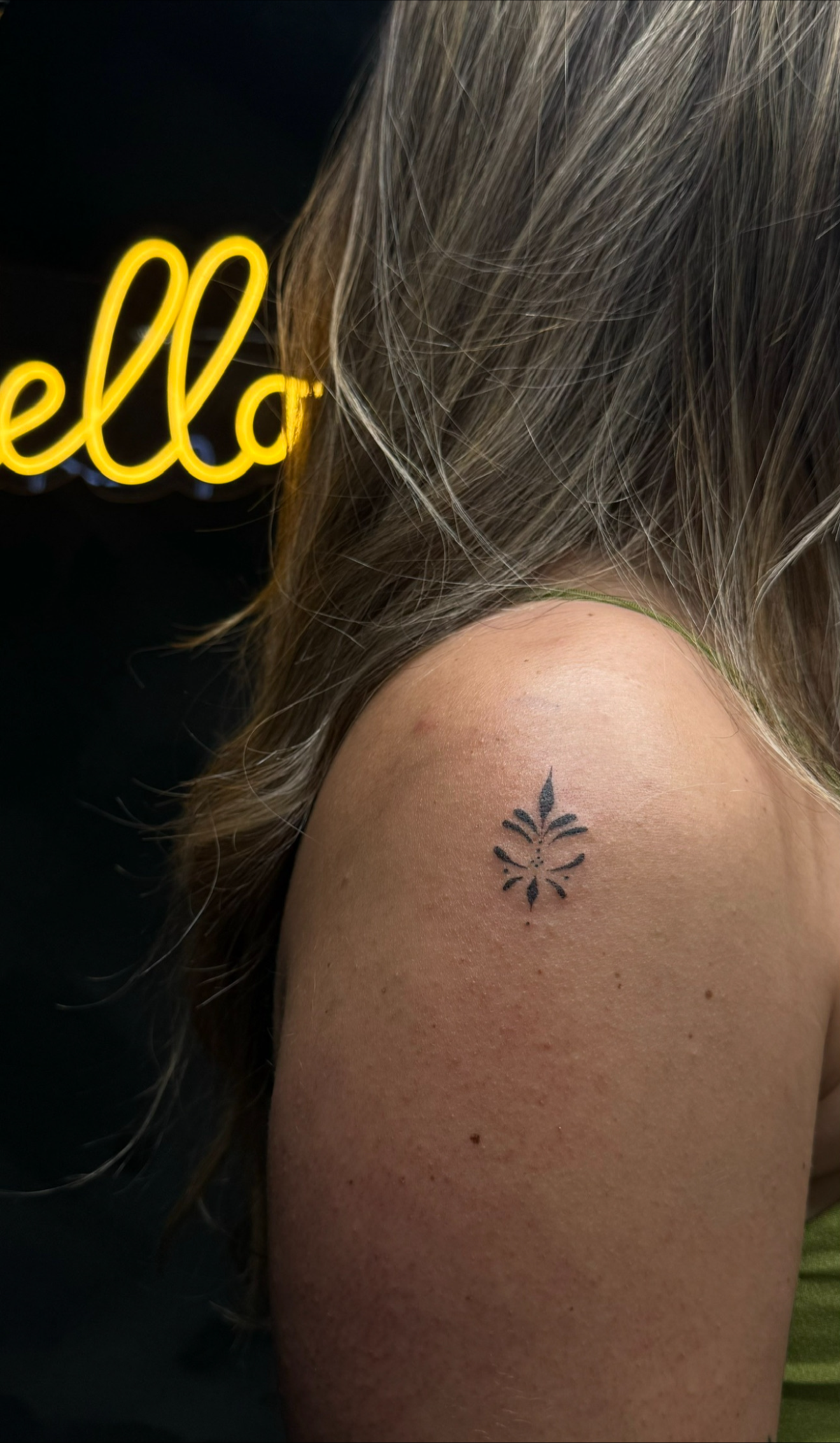 Close-up of a person's shoulder with a small black tattoo of a stylized floral design. In the background, part of a yellow neon sign spelling 'hello' is visible.