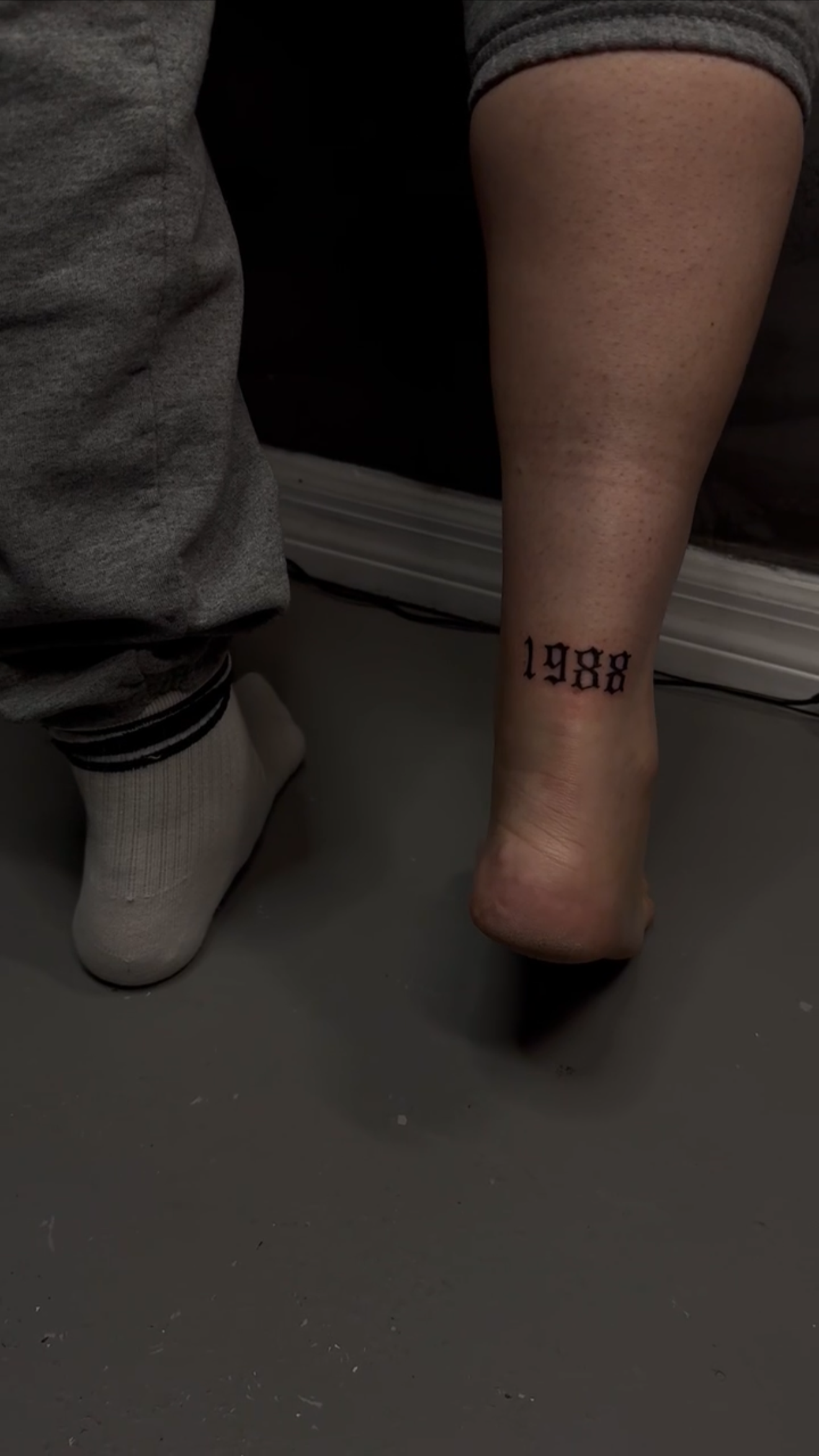 Close-up of a person's ankle with a tattoo reading '1998' in black gothic font, resting on the floor, next to a child's foot wearing a white sock and gray pants.