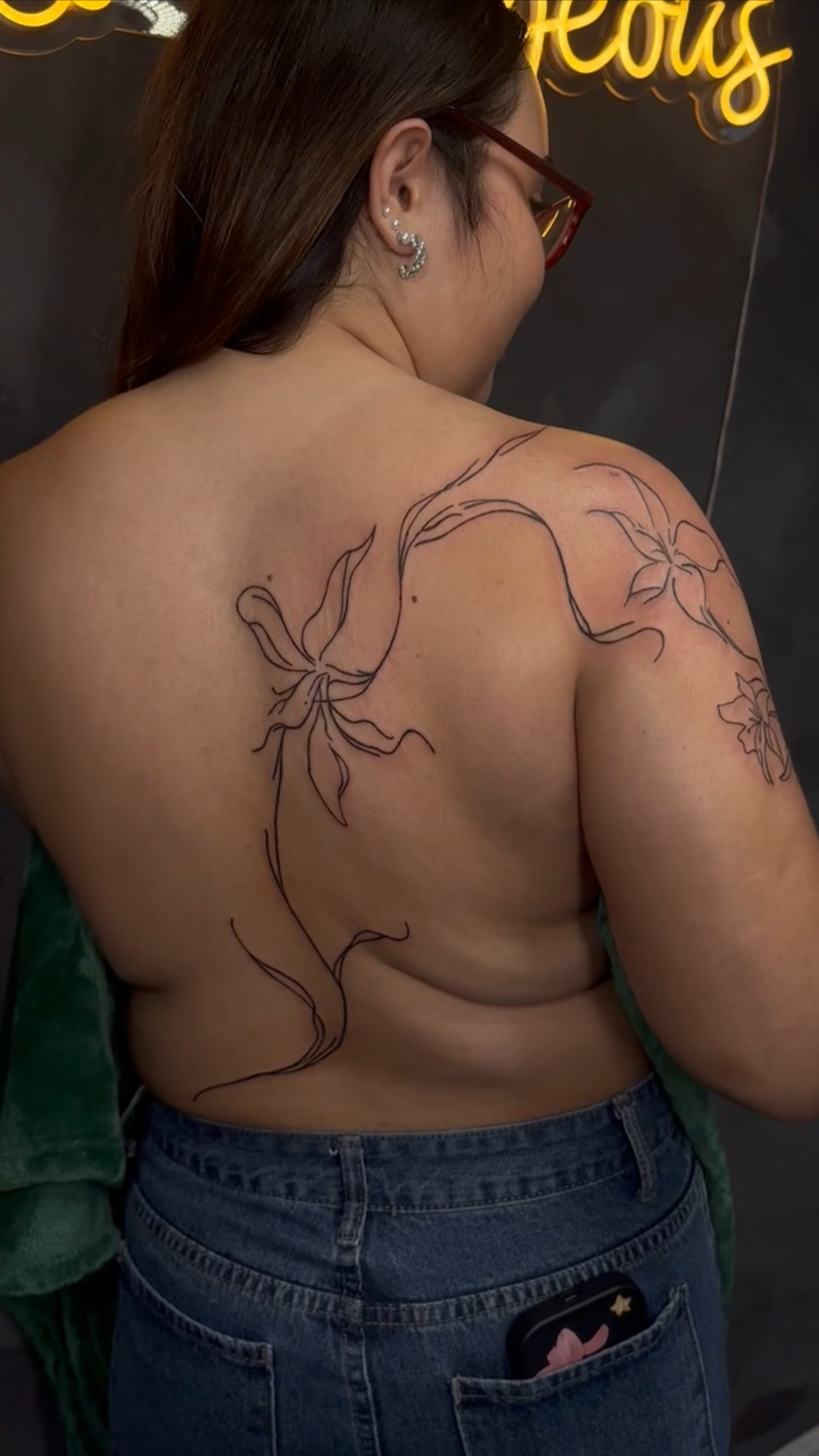 A woman with brown hair and wearing glasses has a tattoo of flowers and leaves on her upper back and shoulder.