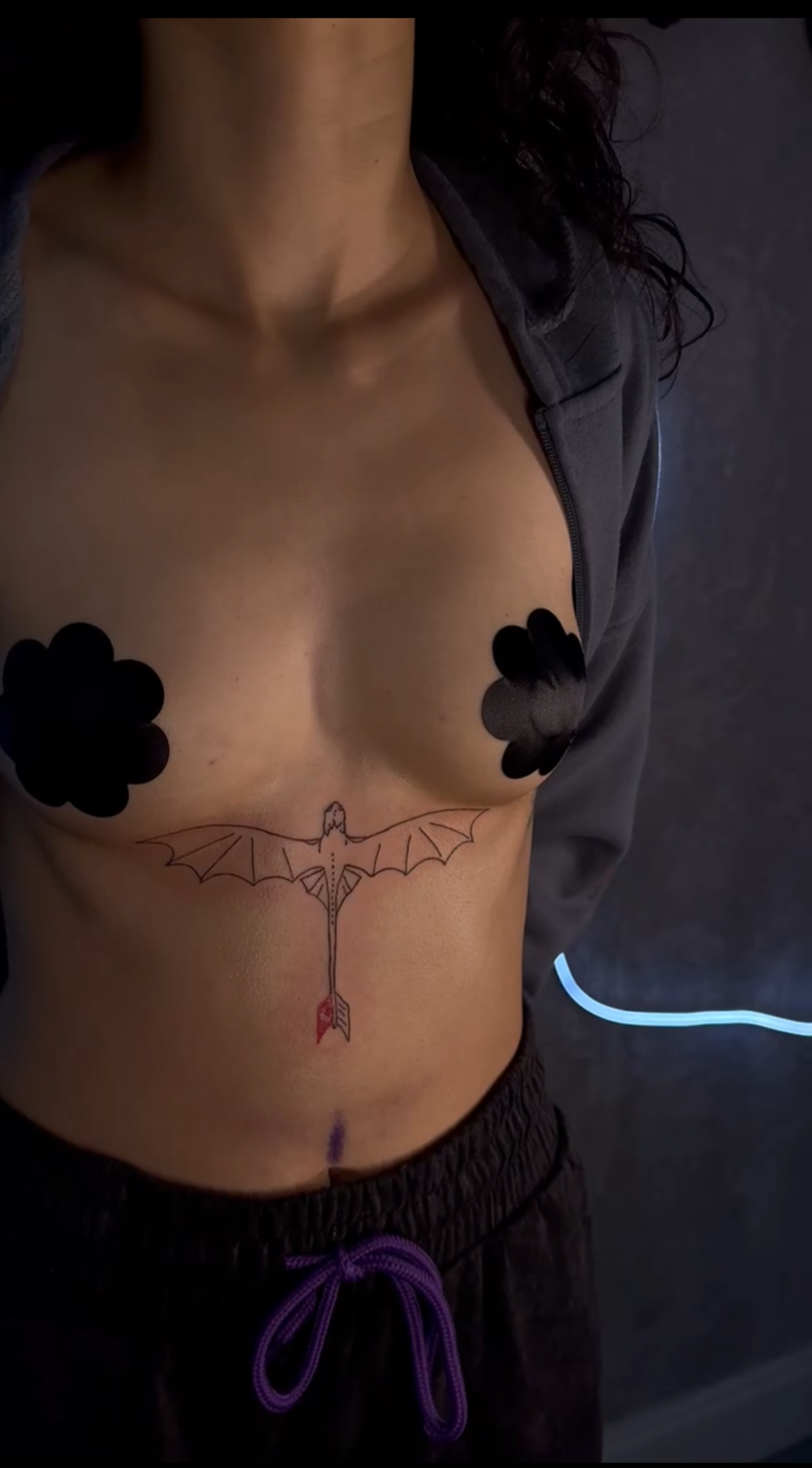 A person with a tattoo of a dragonfly on their side, black flower-shaped pasties covering their nipples, and a zipper tattoo on their lower abdomen, wearing dark pants with purple drawstrings.
