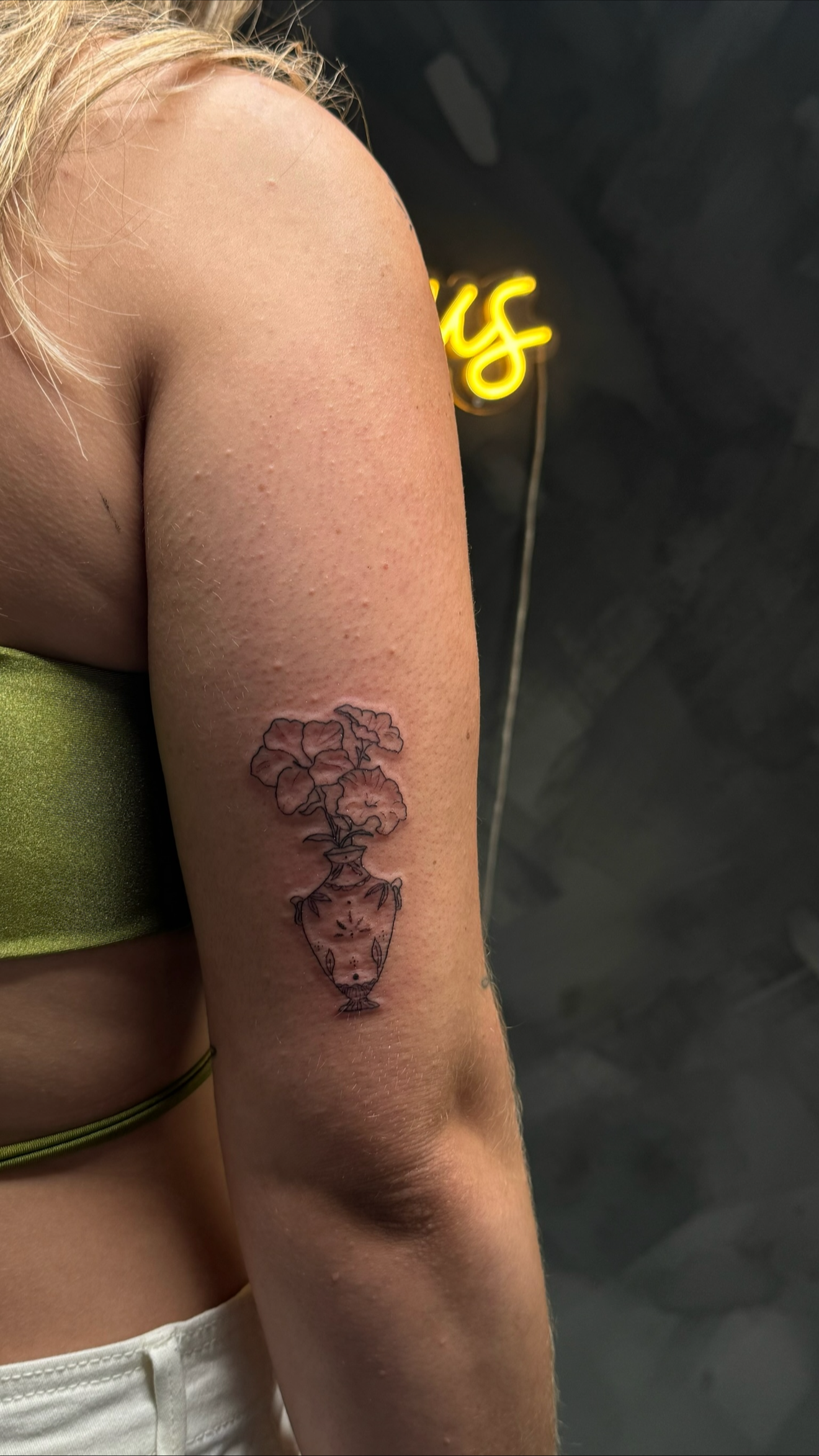 Close-up of a person's upper arm with a tattoo of a vase with flowers, a neon sign in the background, and part of their clothing.