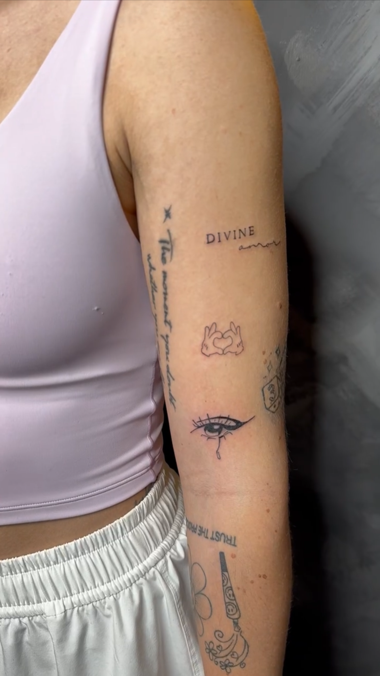 Close-up of a person's arm with various tattoos, wearing a light pink tank top and white pants.
