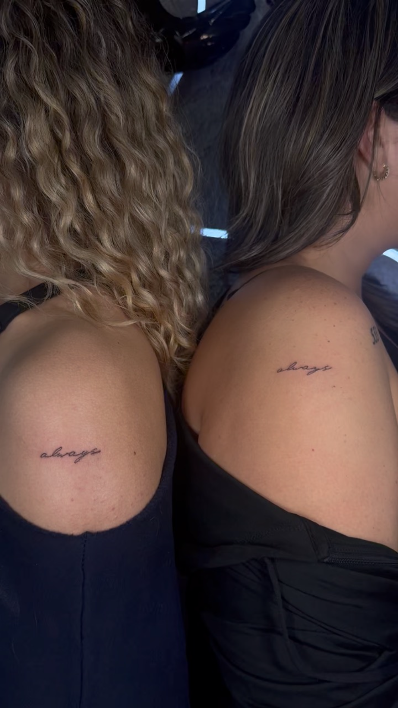 Two women sitting with their backs to the camera, each showing a tattoo on their upper arm that reads "always" in cursive.