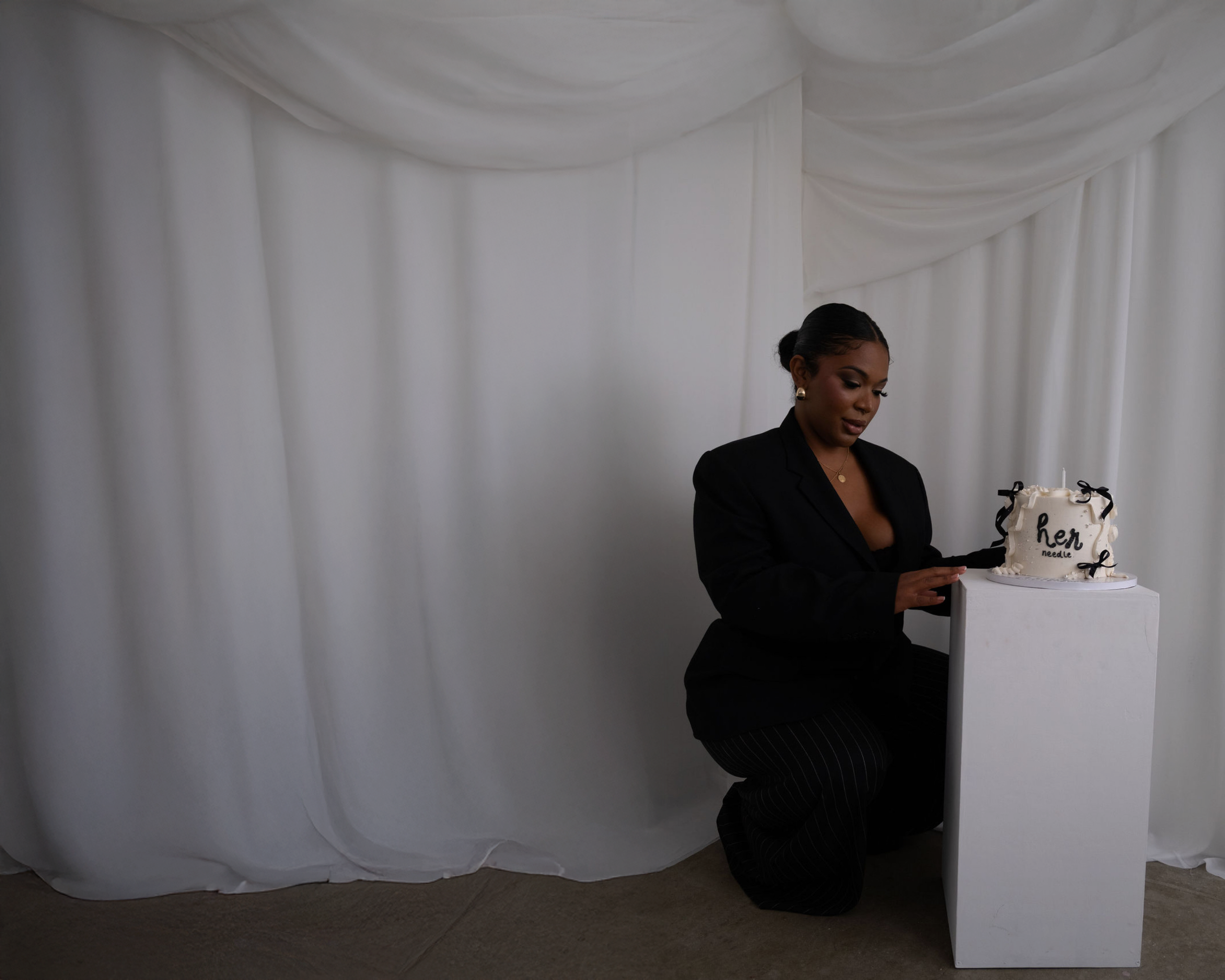 A woman dressed in black with earrings and a necklace, standing in front of a white curtain draped ceiling, holding a wedding cake with black ribbons and a sign that reads "Hen Needle" in black letters on top.