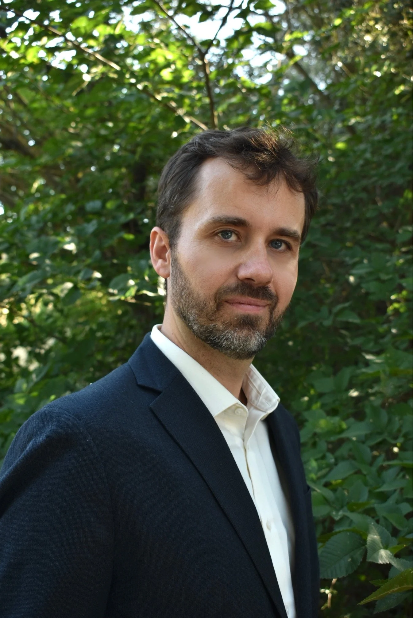 A man dressed in a dark suit and white shirt standing outdoors in front of green foliage, facing slightly to the right with a slight smile.