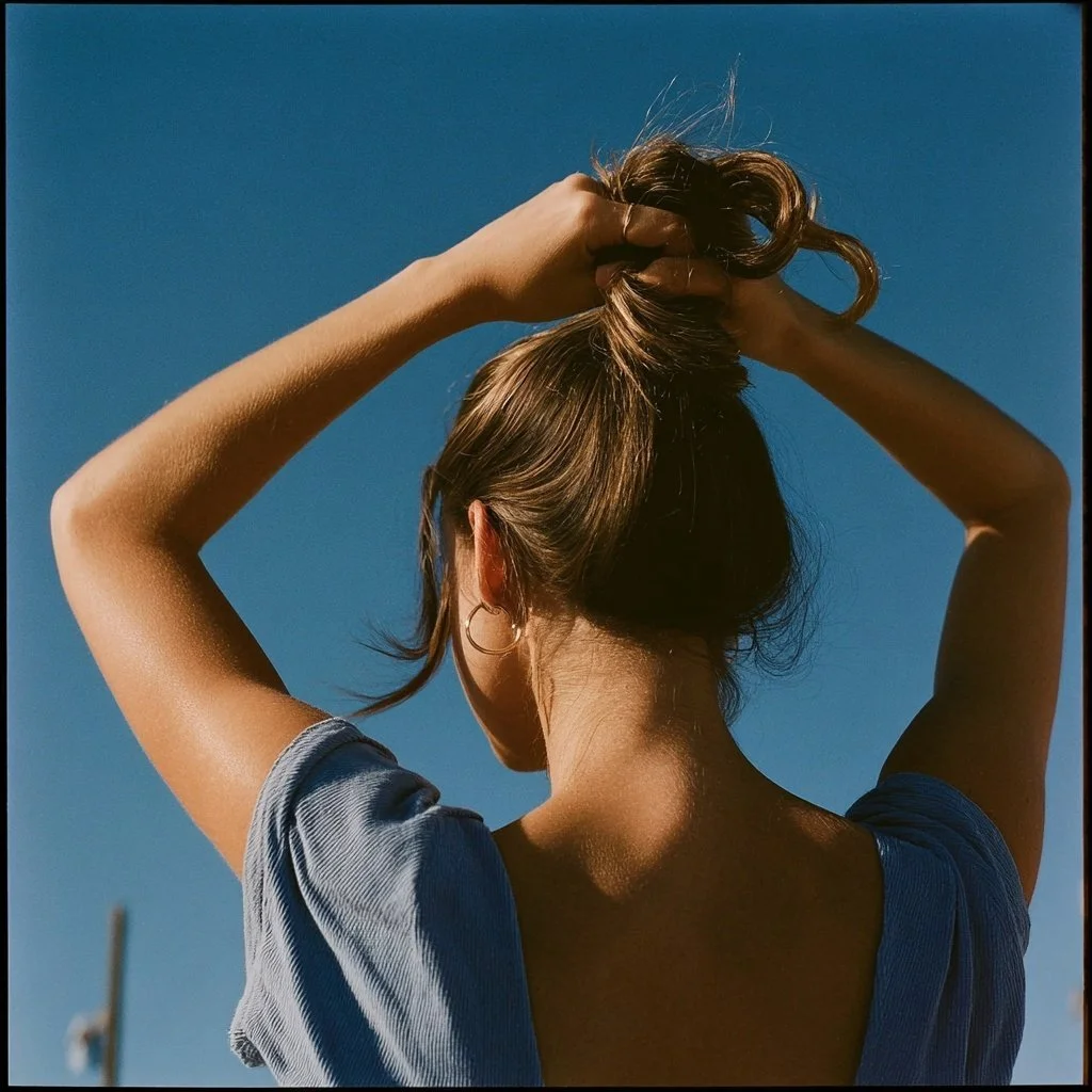 Back of a woman with brown hair in a bun, holding her hair up with both hands against a clear blue sky, wearing a blue shirt with rolled-up sleeves.