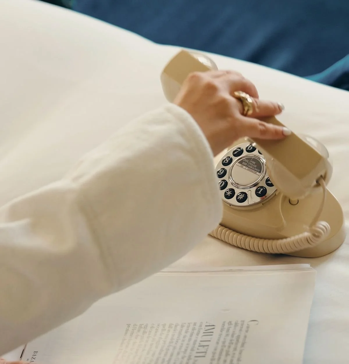 Person holding a beige landline telephone receiver over a white open book on a white table.