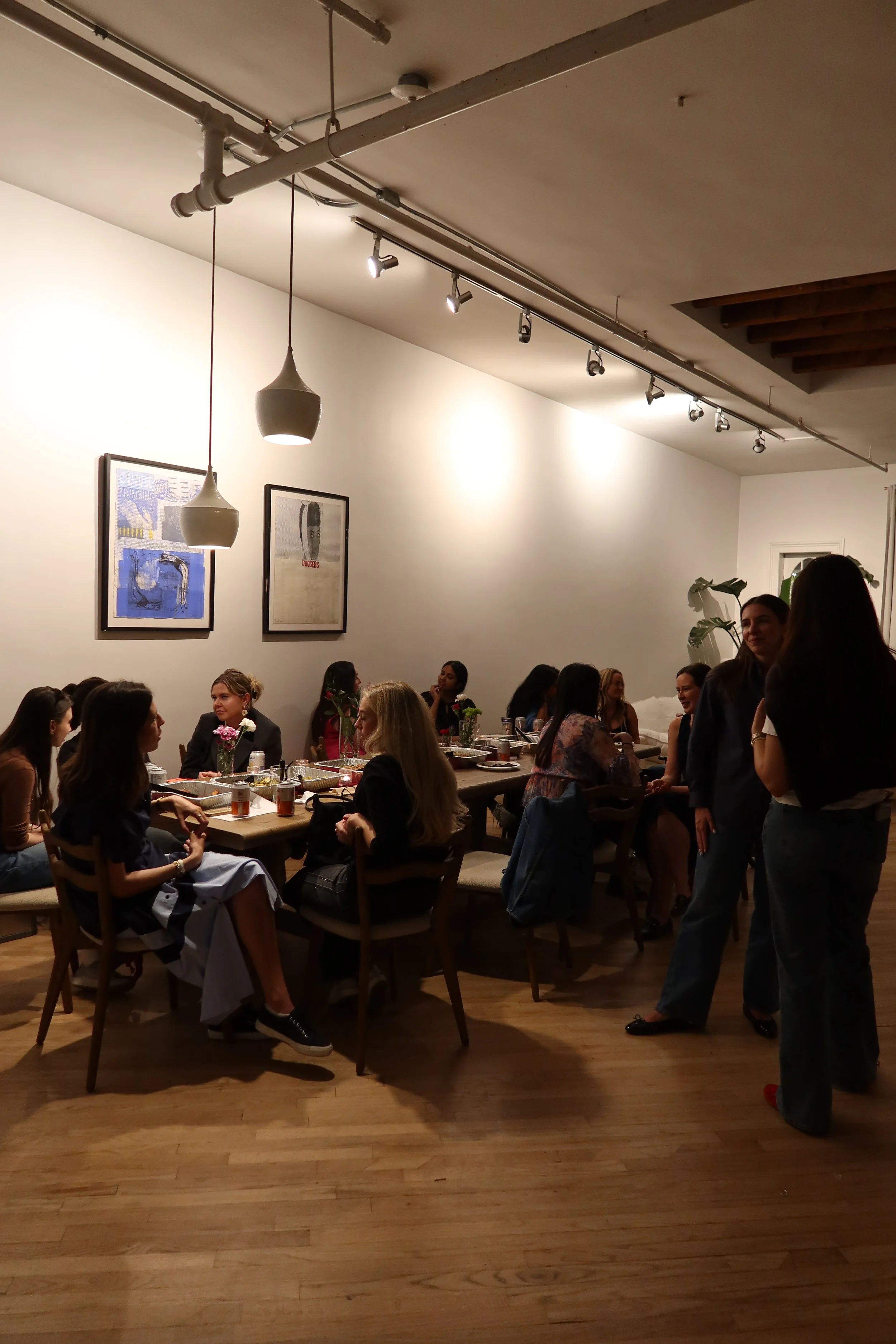 A group of women gathered around a long wooden table in a cozy, well-lit room, engaging in conversation. The room is decorated with framed artwork on the wall, and there are hanging pendant lights and ceiling track lighting. Some women are sitting, and others are standing, appearing to enjoy a social event or casual gathering.