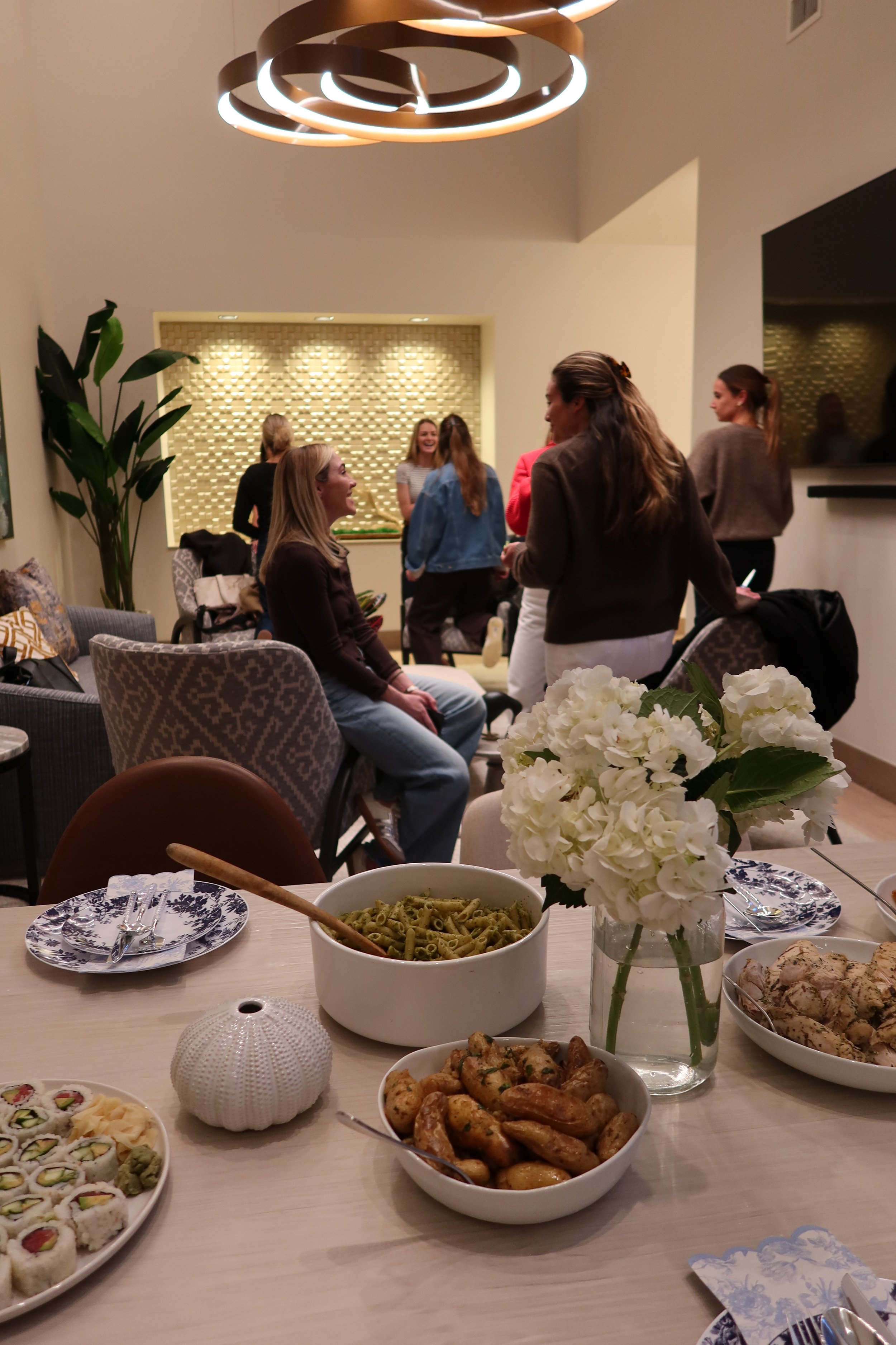 People gathered in a living room, socializing and laughing. There is a modern chandelier hanging from the ceiling. A table in the foreground has sushi, pasta, and other dishes, along with a vase of white hydrangeas.