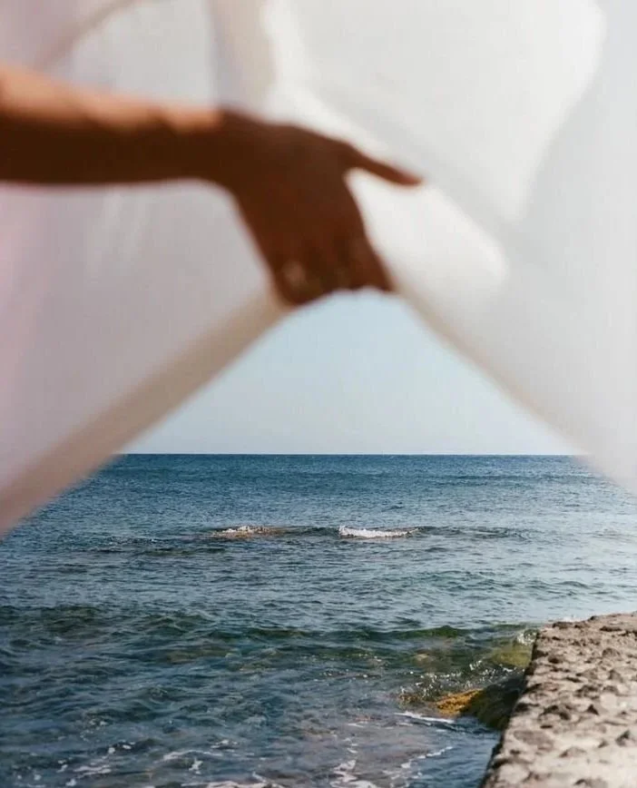 A person holding a white fabric or cloth up to frame a view of the ocean and shoreline with gentle waves and rocks.