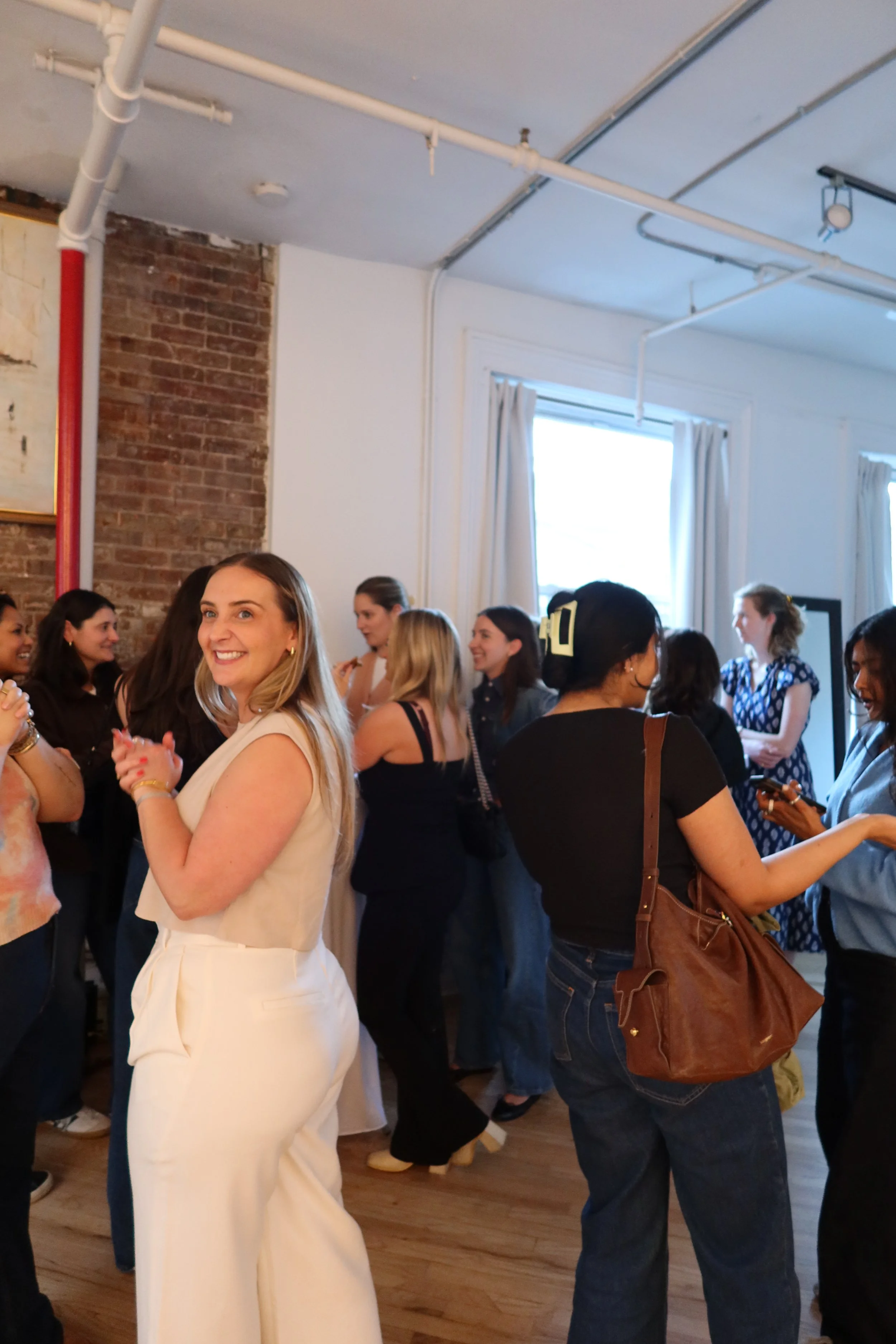 A group of people socializing and mingling in a room with white walls, large windows with white curtains, and a brick accent wall. The people are smiling, some facing the camera, and are dressed in casual to semi-formal attire.