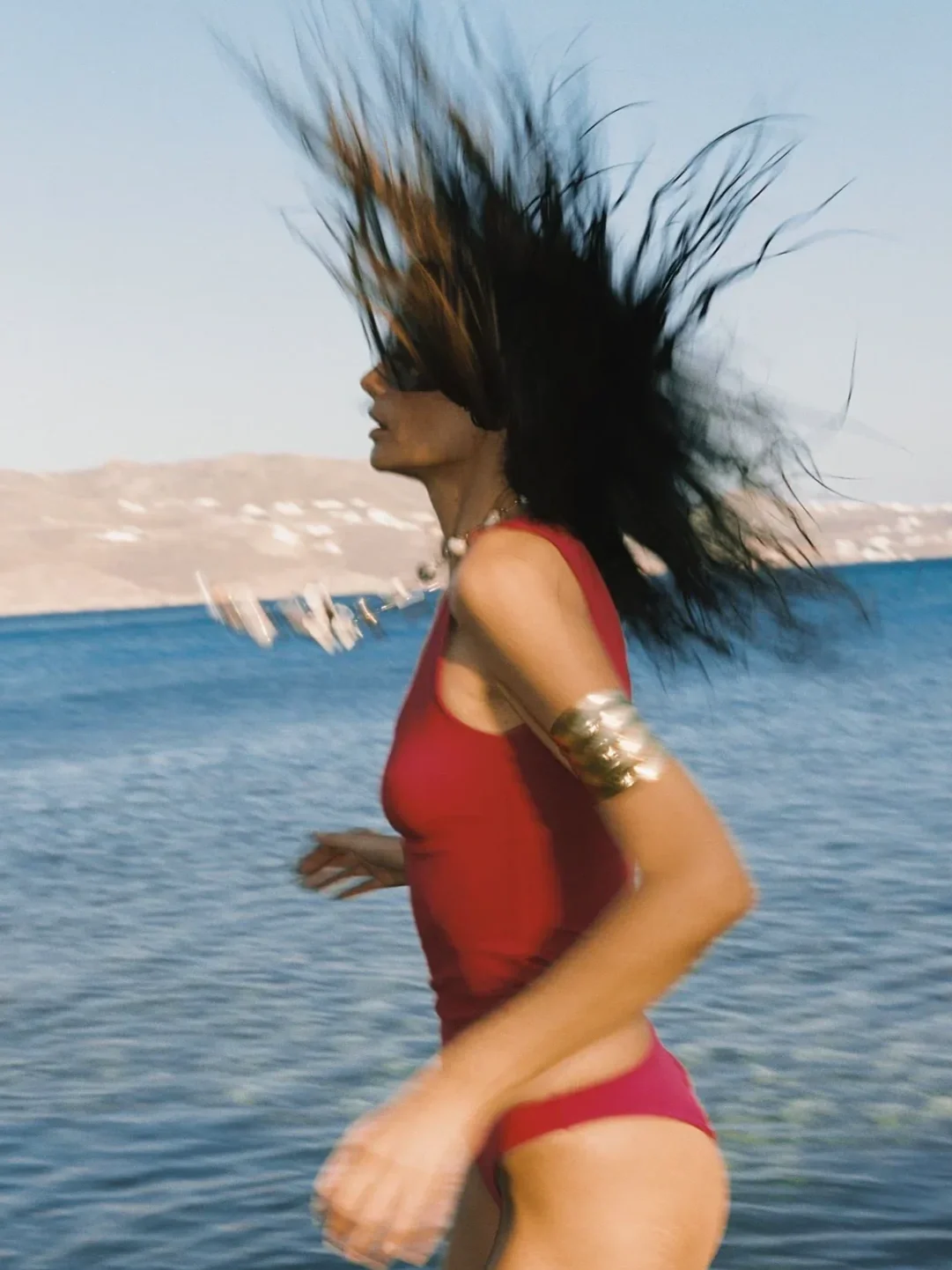 Woman running near water with her hair moving in the wind, wearing a red swimwear and accessories, with boats and mountains in the background.