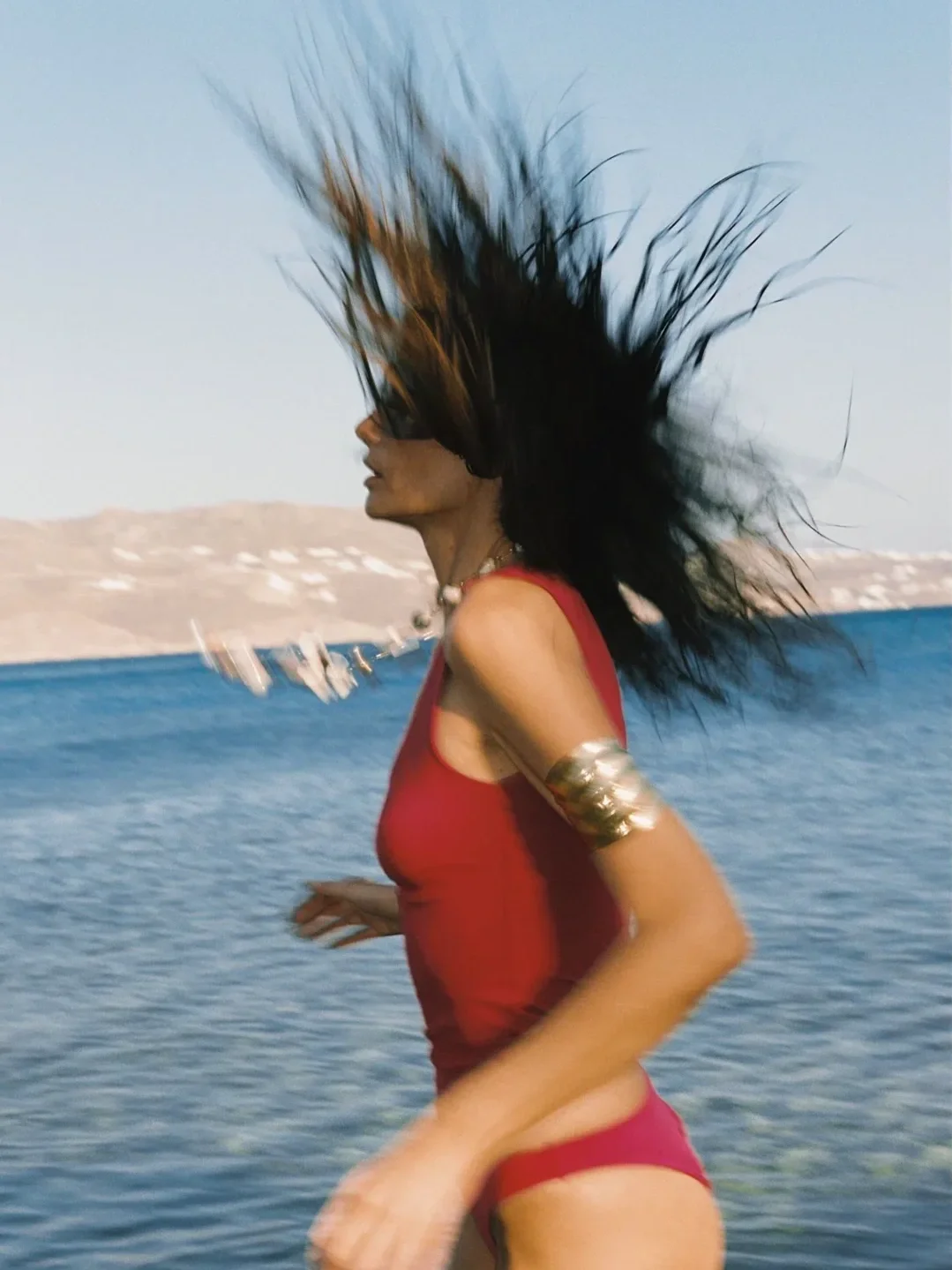 A woman with long dark hair flowing in the wind, wearing a red swimsuit and metallic accessories, running by the water with hills and sailboats in the background.