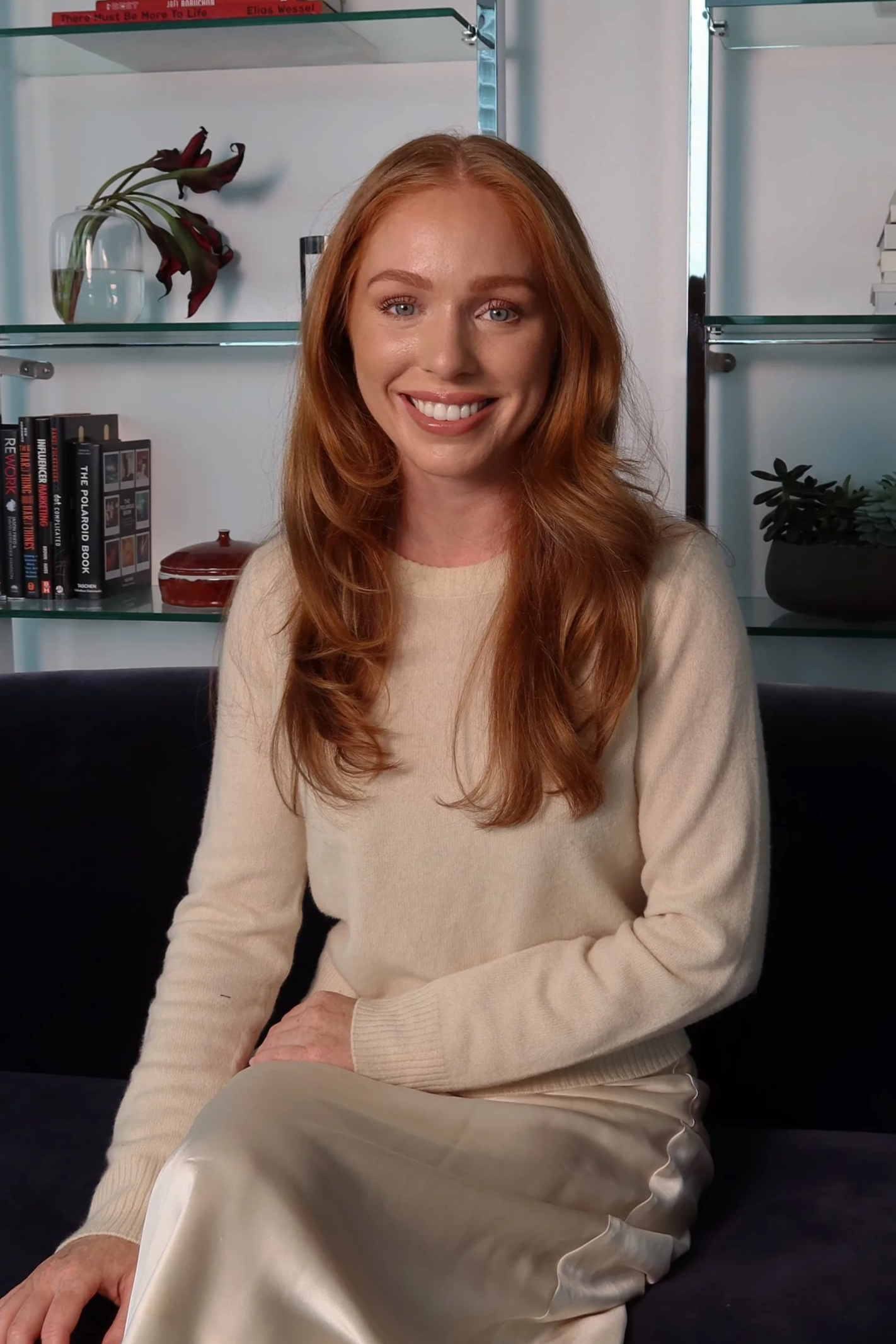 A smiling woman with long red hair sitting on a black cushioned sofa in front of a bookshelf with books and decorative items in a modern room.