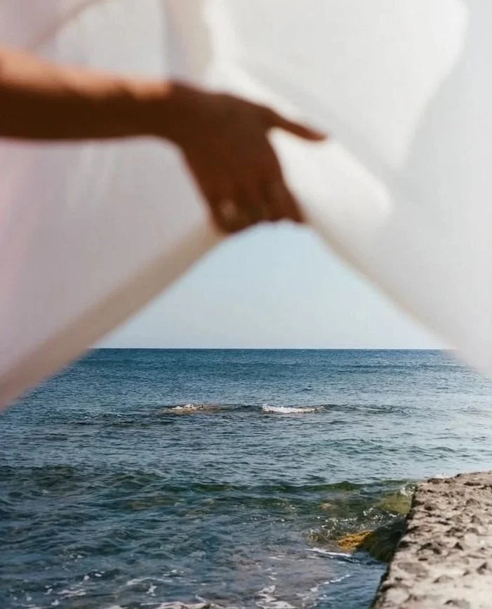 A person holding a book open with the ocean and a rocky shoreline visible through the pages.