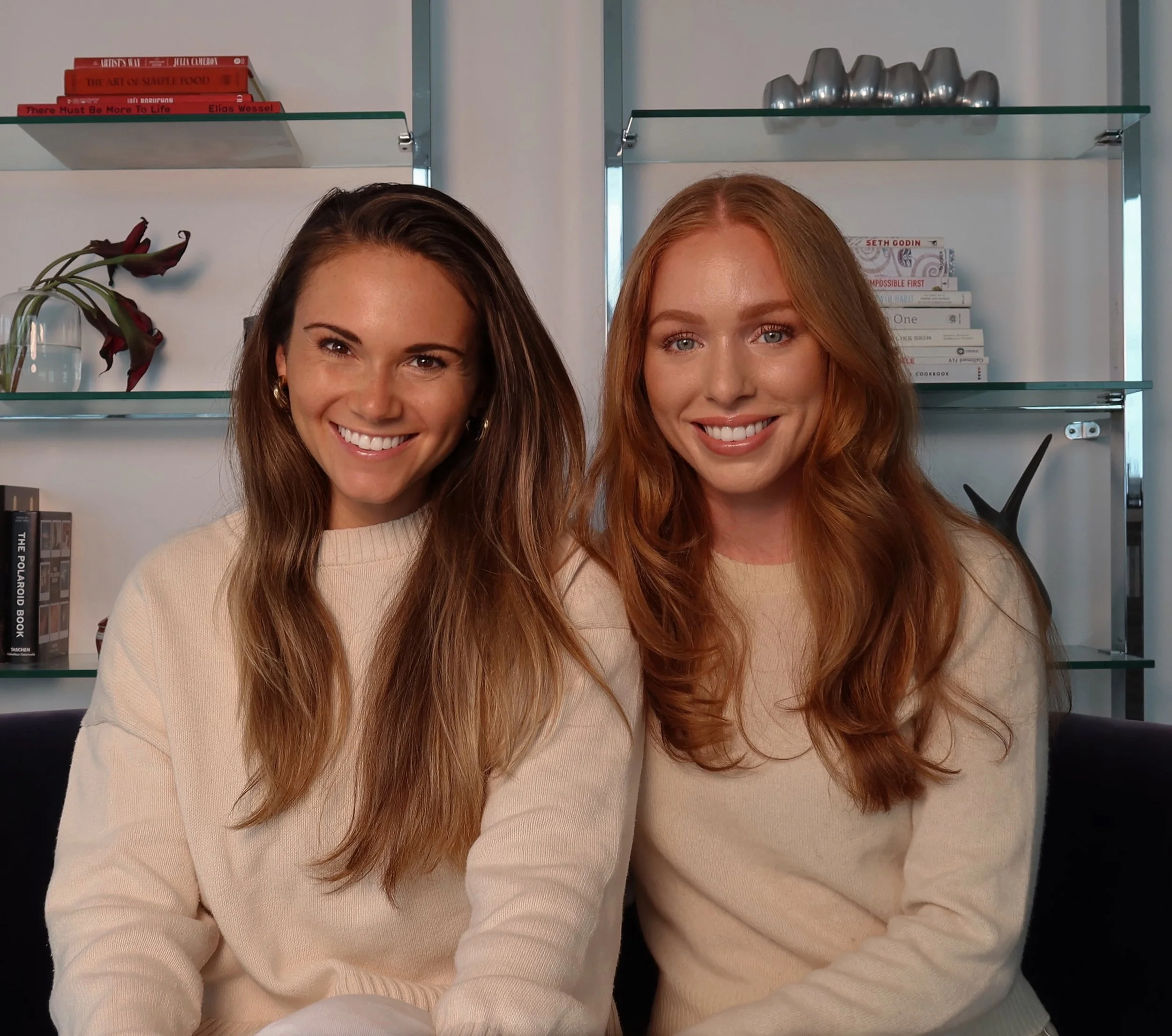 Two women smiling, standing close together in front of glass shelves with books and decorative objects in a modern interior.