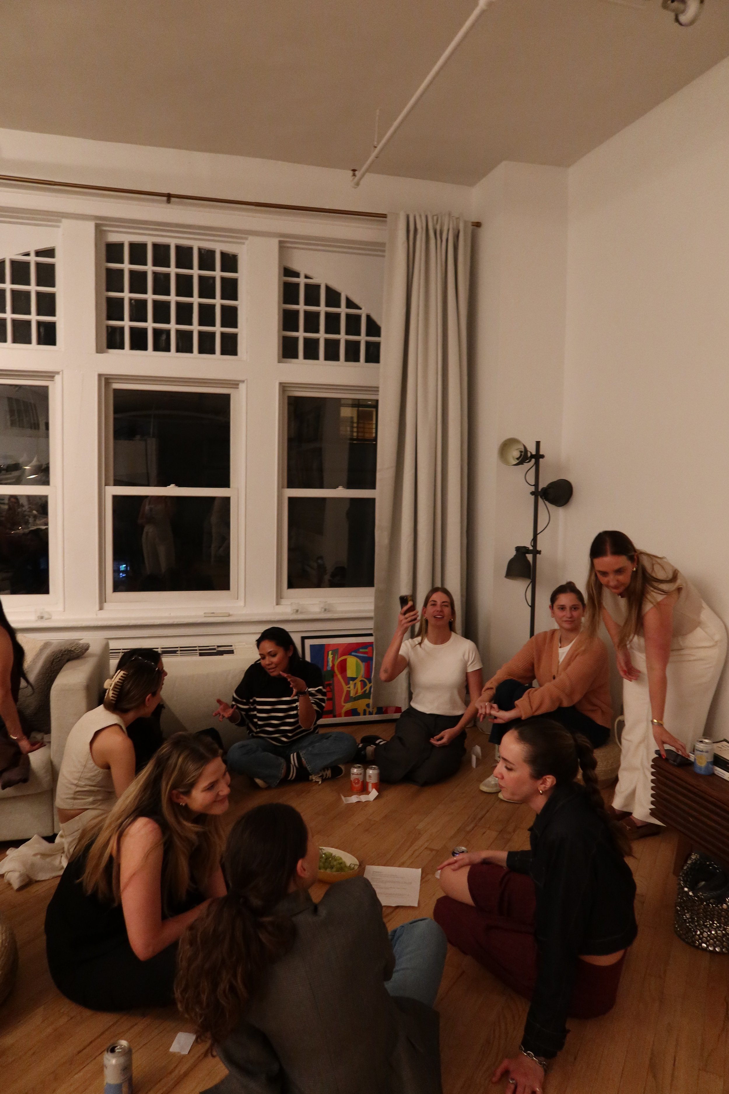 A group of women gathered in a living room, sitting on the floor and on a sofa, engaging in conversation and smiling. Some are taking pictures or videos, with various drinks and snacks around.