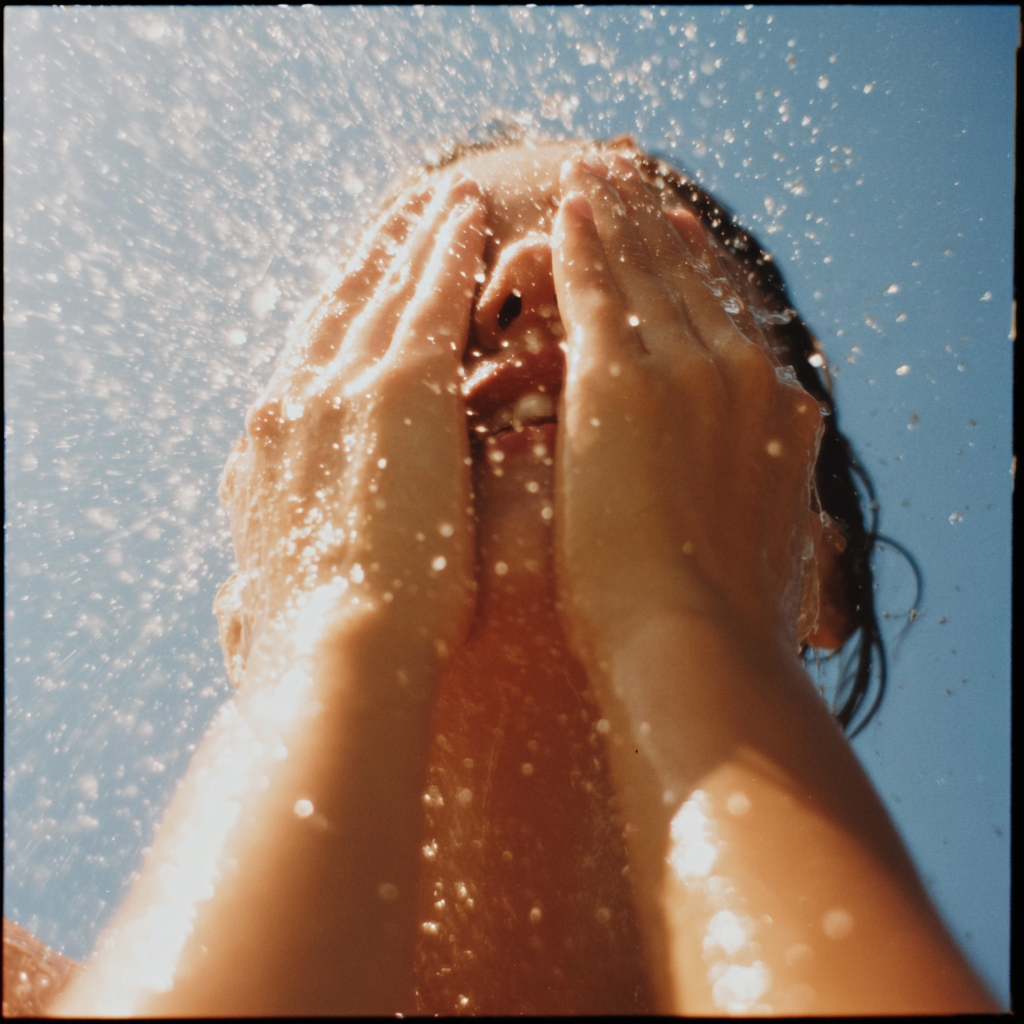 A woman is splashing water on her face under the sun, with water droplets surrounding her and a clear blue sky in the background.