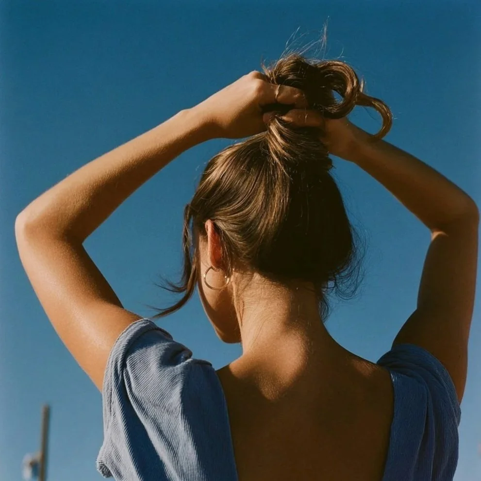 A woman with brown hair, wearing a blue shirt, is standing outdoors against a clear blue sky. She is holding her hair up with both hands.