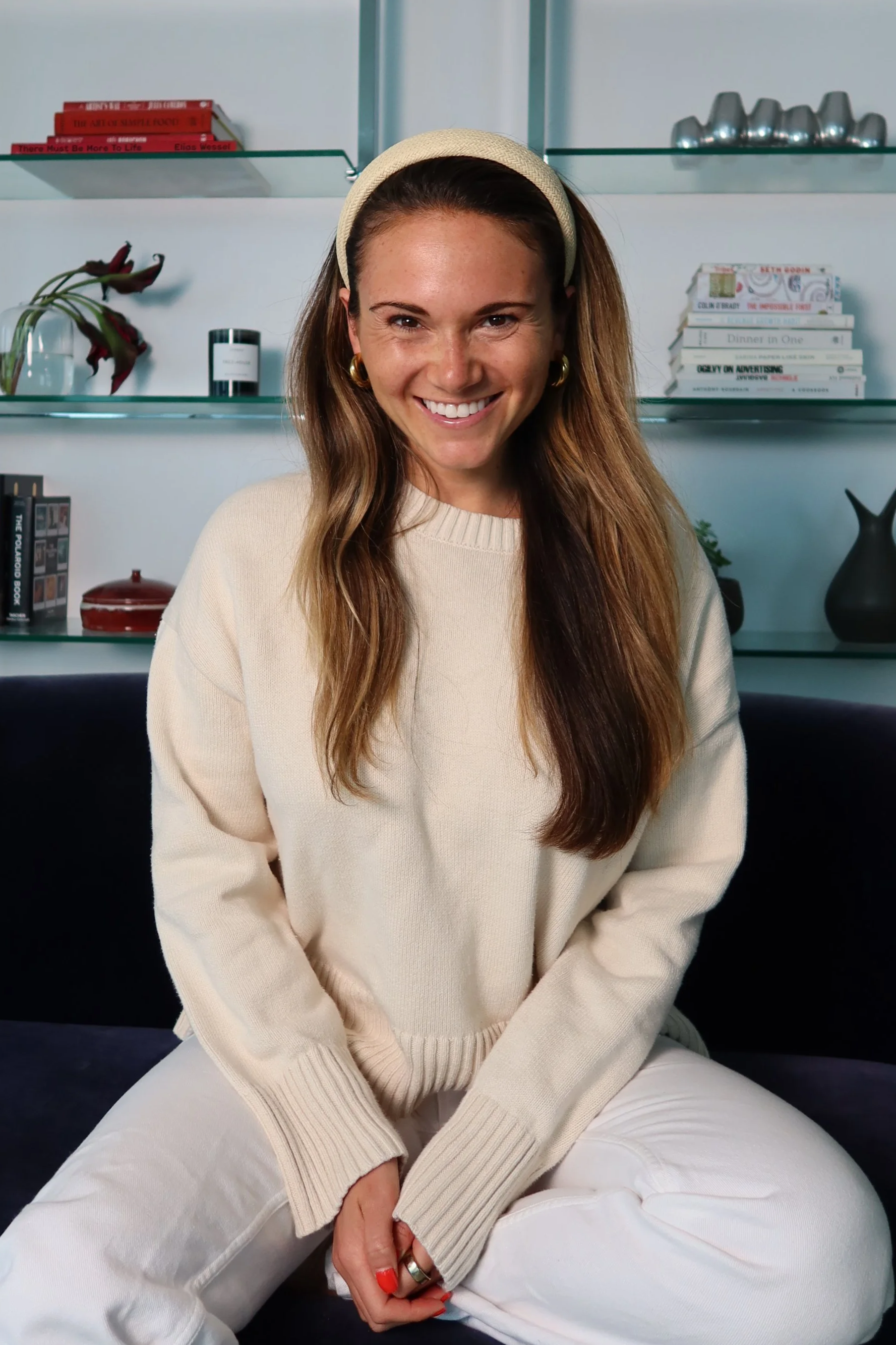A woman with long brown hair, wearing a beige headband and beige sweater, sitting on a black surface in front of glass shelves with books and decorative items.