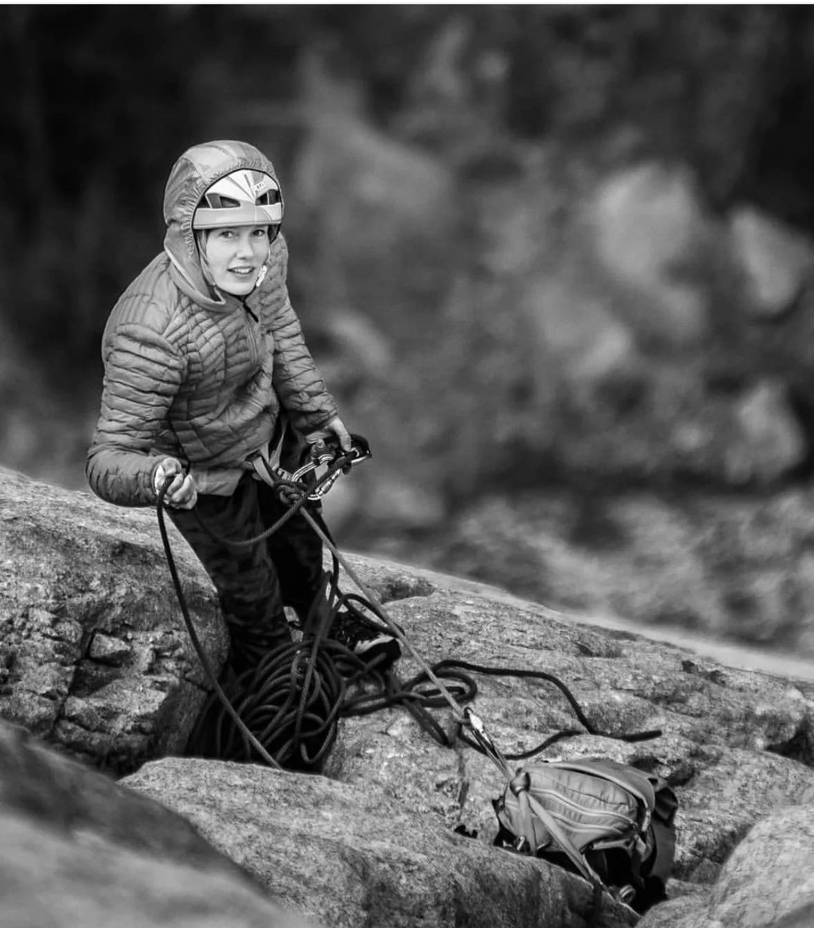 A person wearing a helmet and outdoor jacket, sitting on rocks during rock climbing or canyoneering, with climbing gear and rope on the ground.