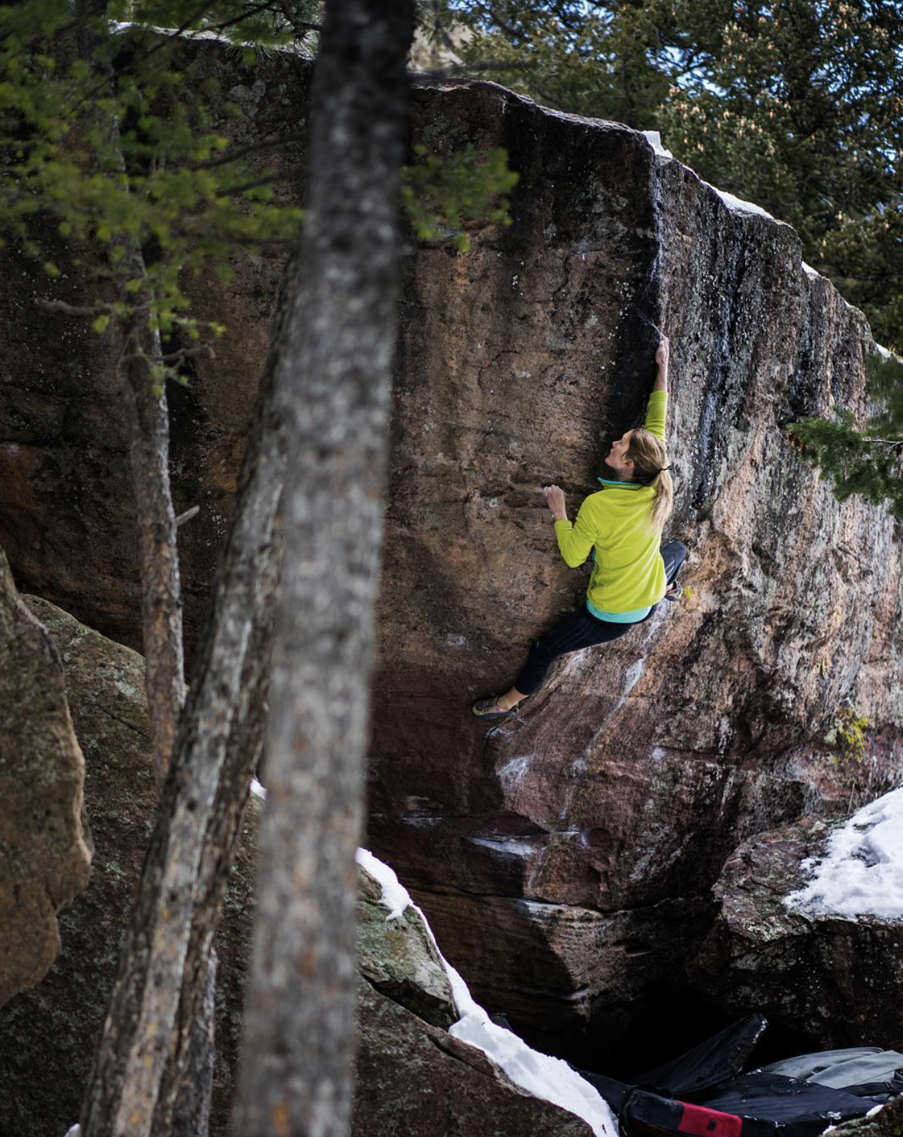 A woman climbing a steep rock face outdoors surrounded by trees and snow.