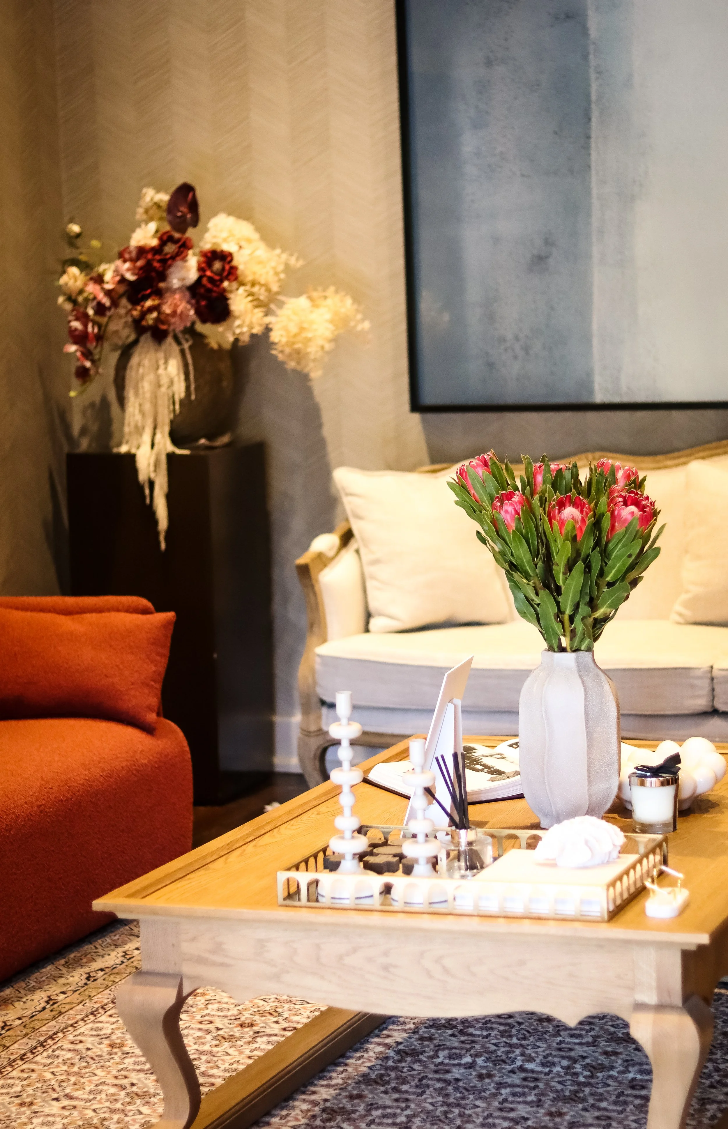 A cozy living room with a wooden coffee table holding a vase of pink flowers, a chess set, and decorative items. In the background, there is a cream-colored sofa with pillows, a dark side table with a floral arrangement, and a large wall art piece. The room has a patterned rug and warm lighting.