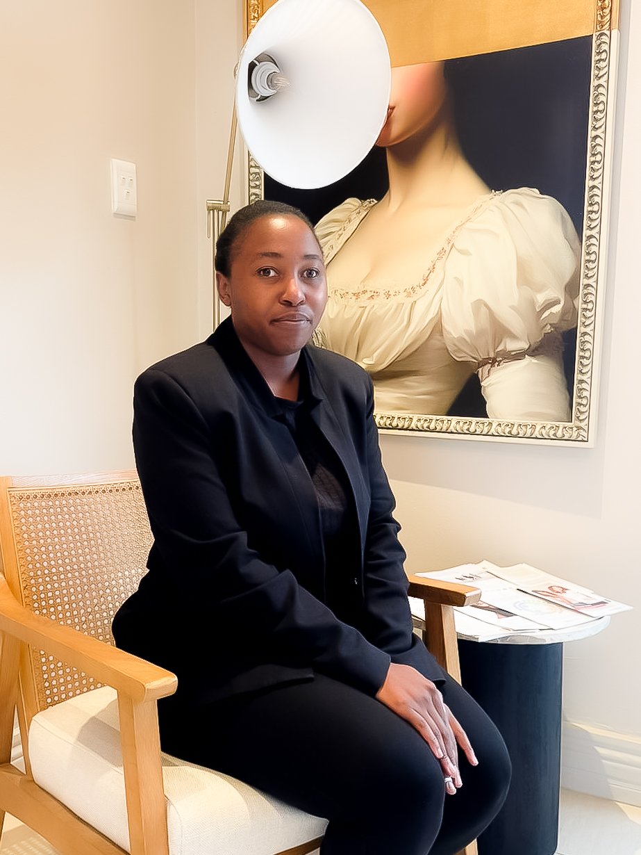 A woman sitting on a wooden chair with a beige cushioned seat in an art gallery or museum. She is wearing a black jacket and black pants. Behind her is a large framed painting of a woman in a cream-colored dress with puffed sleeves, holding a large white circular object in front of her face, obscuring her features. There are papers and magazines on a small black table next to her.
