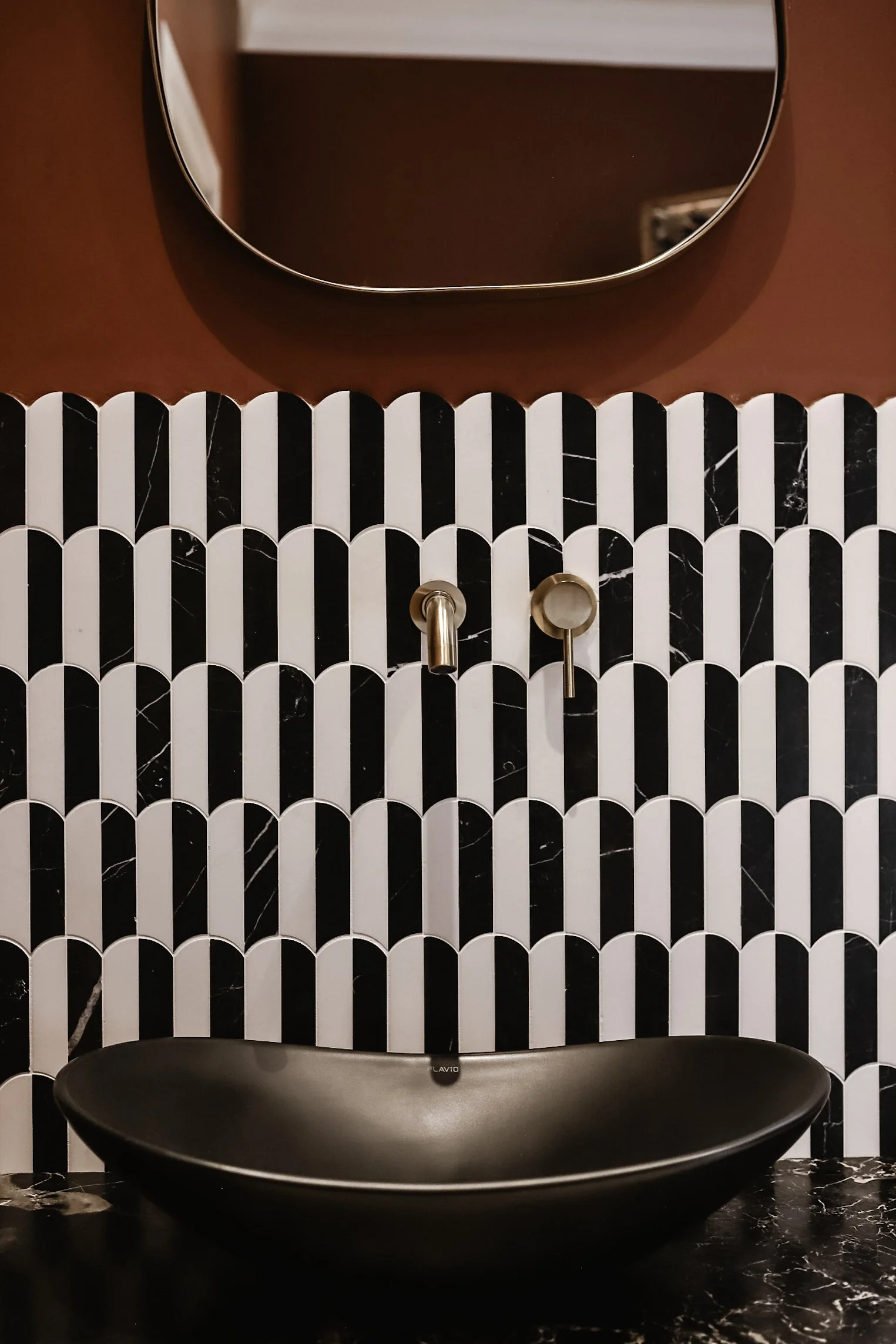 A modern black basin sink with a metallic faucet in a bathroom with black and white patterned tiles and a round mirror above.