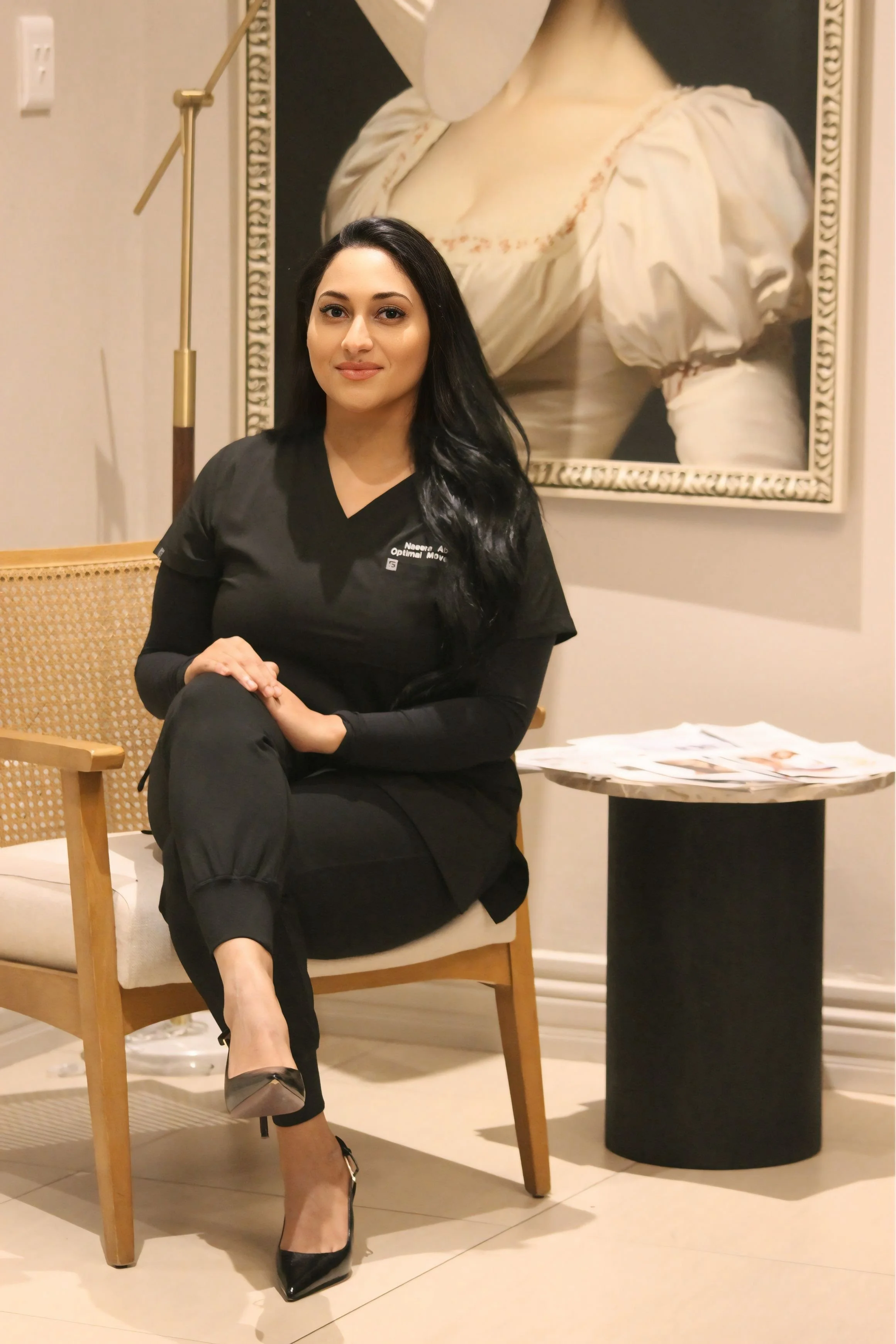 A woman with long black hair and light skin tone sitting on a wooden chair with a woven backrest. She is wearing black scrubs, black high heels, and has one leg crossed over the other. Behind her, there's a large framed painting of a woman in a white dress with puffed sleeves. To her right, there's a black round table with folded papers on top and a tall, black trash bin. The setting appears to be indoors with beige walls and tiled flooring.