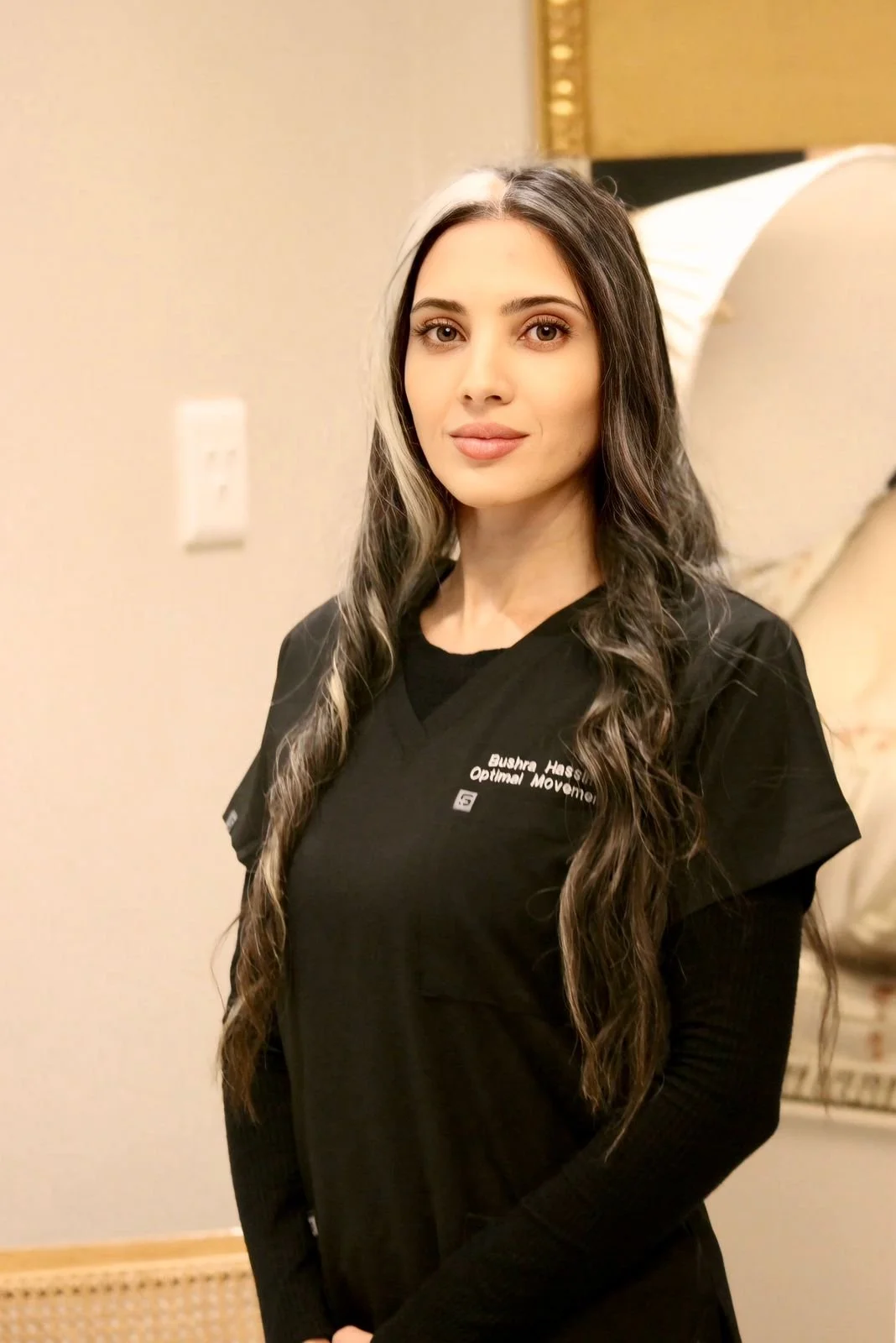 A woman with long, wavy dark hair with a streak of white, wearing a black medical scrub top with white text, standing in a room with beige walls, a white light switch, and a bed in the background.