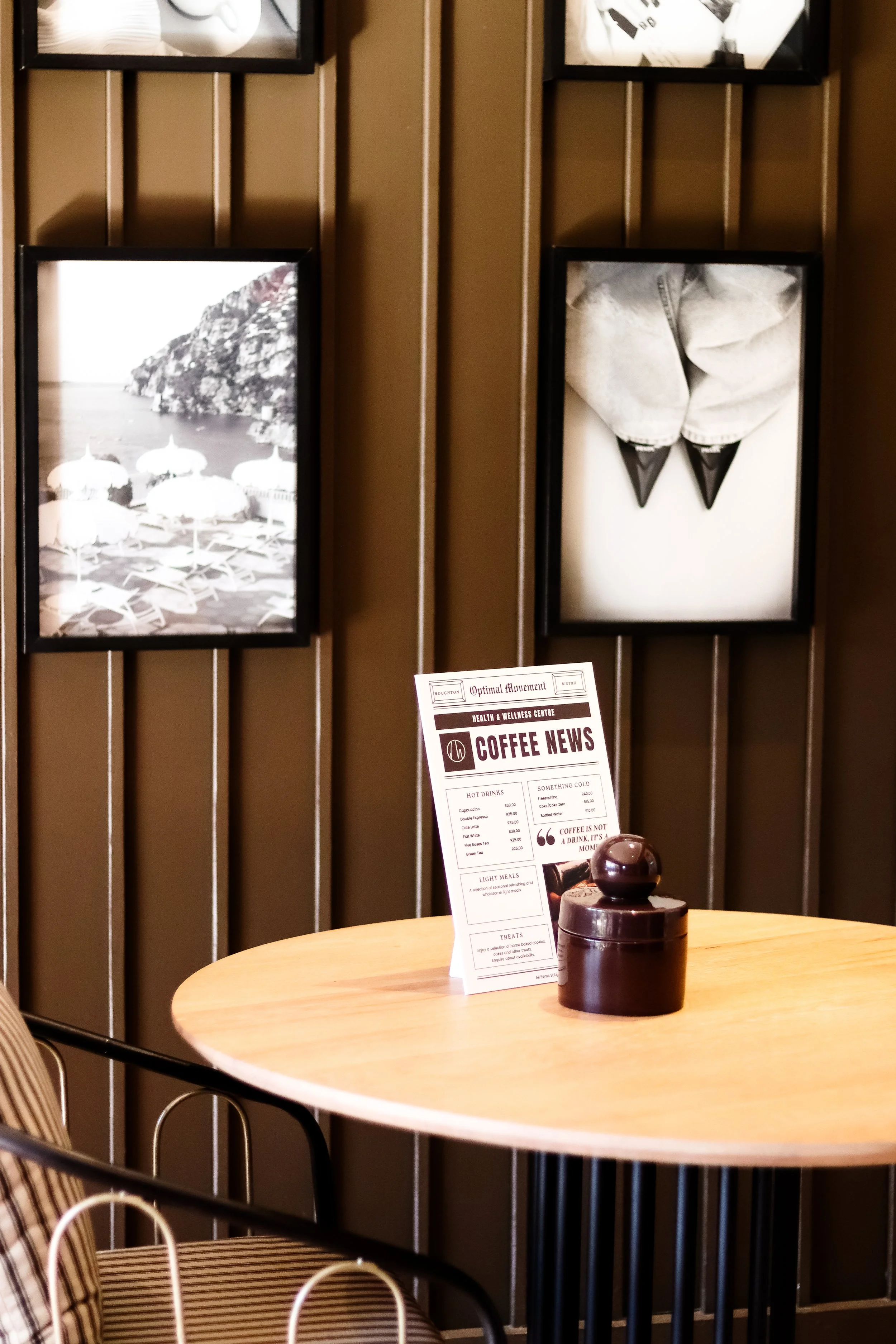 Indoor scene of a cafe or restaurant with a round wooden table, a menu on the table, dark-colored container, and black framed photos on a brown wall.