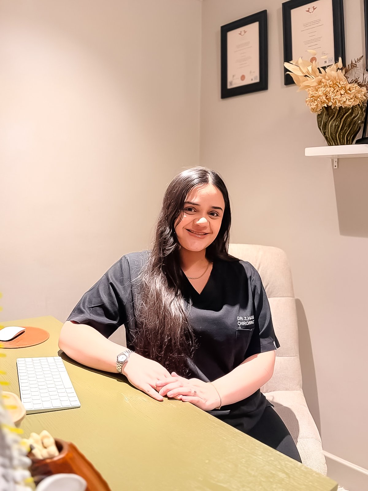 A smiling woman with long dark hair sitting at a desk in a medical office, wearing a black scrubs with 'Dr. T. Husi' and 'Chiropractic' embroidered on it, with framed certificates on the wall behind her and a vase of dried flowers on a shelf to the right.