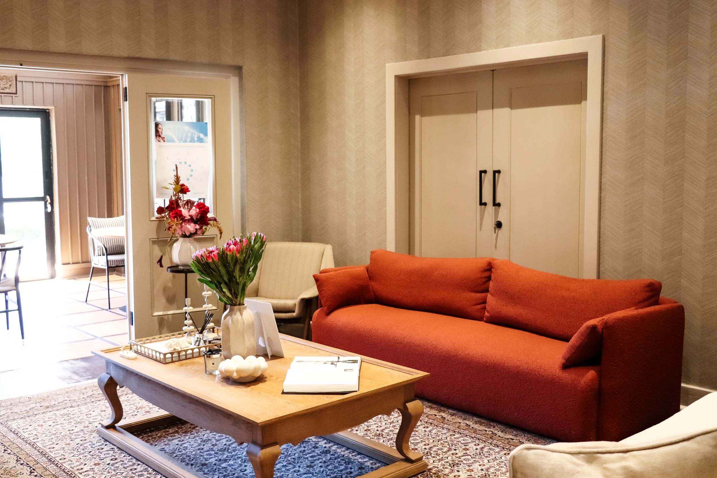 A cozy living room with a wooden coffee table, red sofa, cream-armchair, vase with pink flowers, and decorative items on the table. There is a patterned rug on the floor, and in the background, a door, framed artwork, and a window or door with natural light.