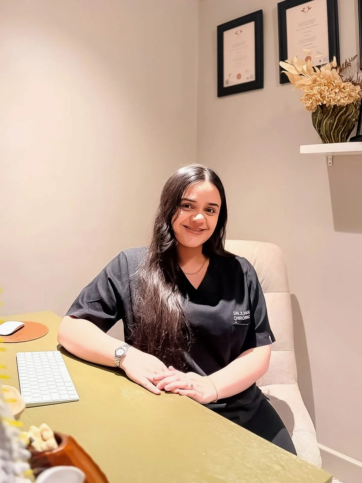 A woman with long dark hair, wearing a black medical uniform with a name tag, sitting at a desk in an office or clinic. She is smiling and has a beige chair behind her. There are framed certificates on the wall behind her and a decorative vase with flowers on a shelf.