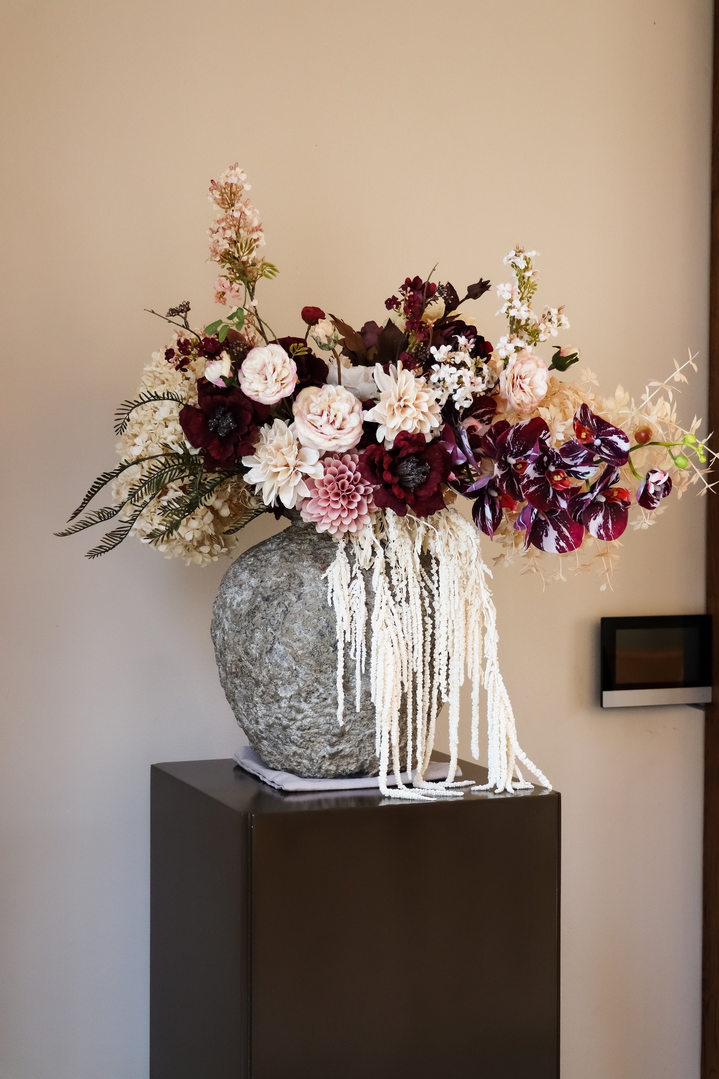 A large textured stone vase with a cascading white beaded decoration spilling over the edge, filled with a bouquet of mixed flowers including pink roses, white dahlias, dark burgundy blooms, and purple orchids, positioned on a black pedestal against a plain beige wall.