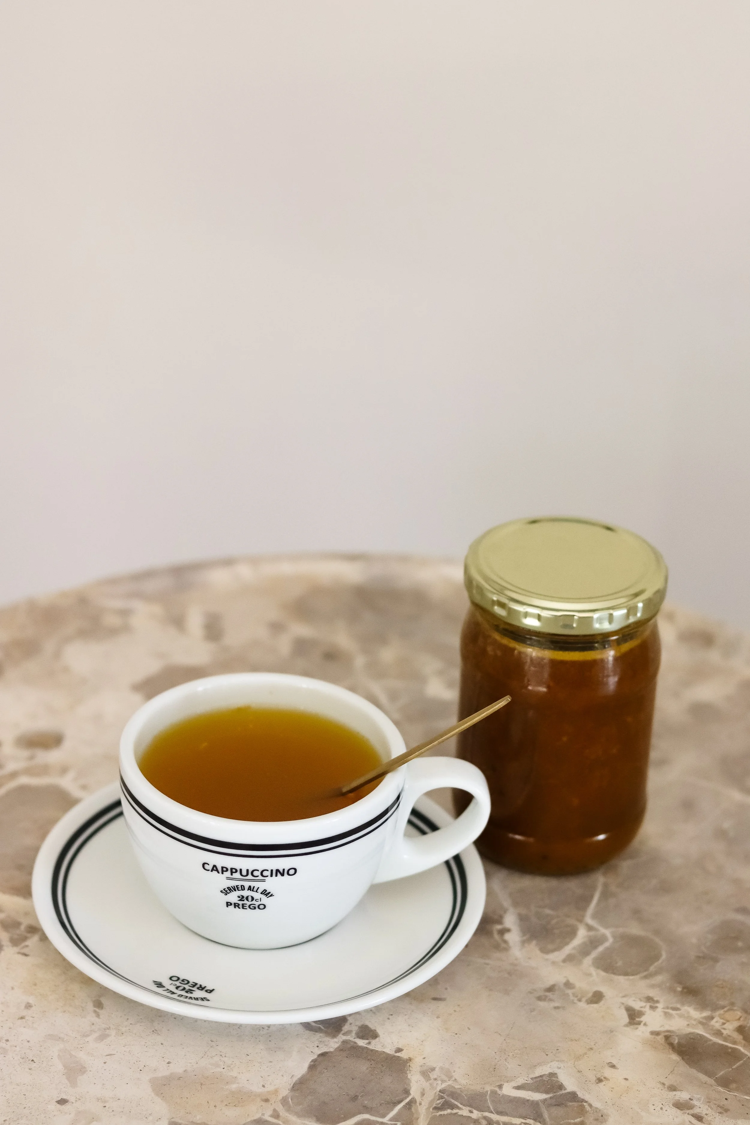 A white cup of tea on a saucer with black stripes and the word 'CAPPUCCINO' written on it. There is a spoon resting inside the cup. In the background, there is a jar of honey and a marble countertop.