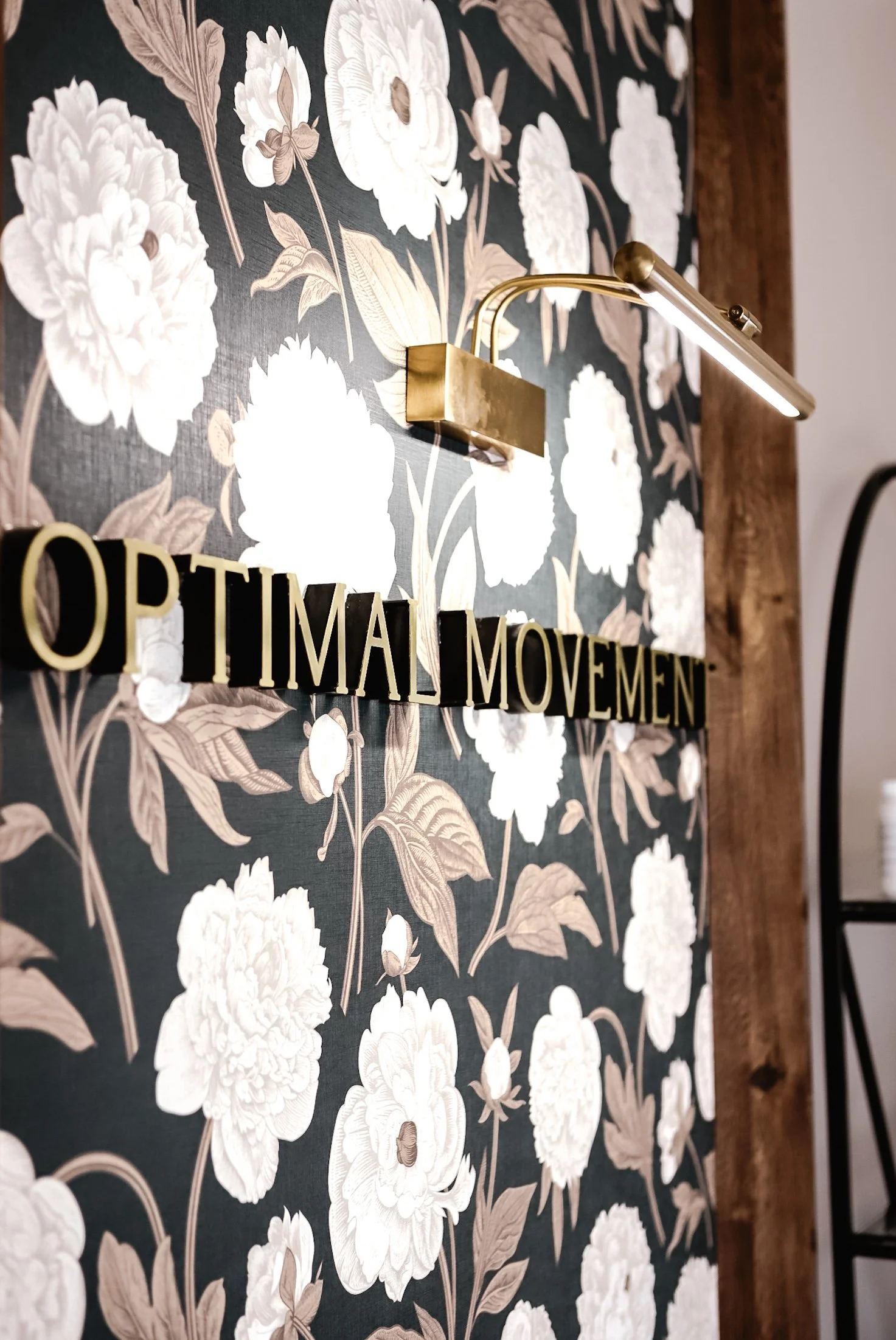 Interior wall with black and cream floral wallpaper, gold wall-mounted light fixture, and a decorative sign with black and beige letters reading 'OPTIMISTIC MOVEMENT'.